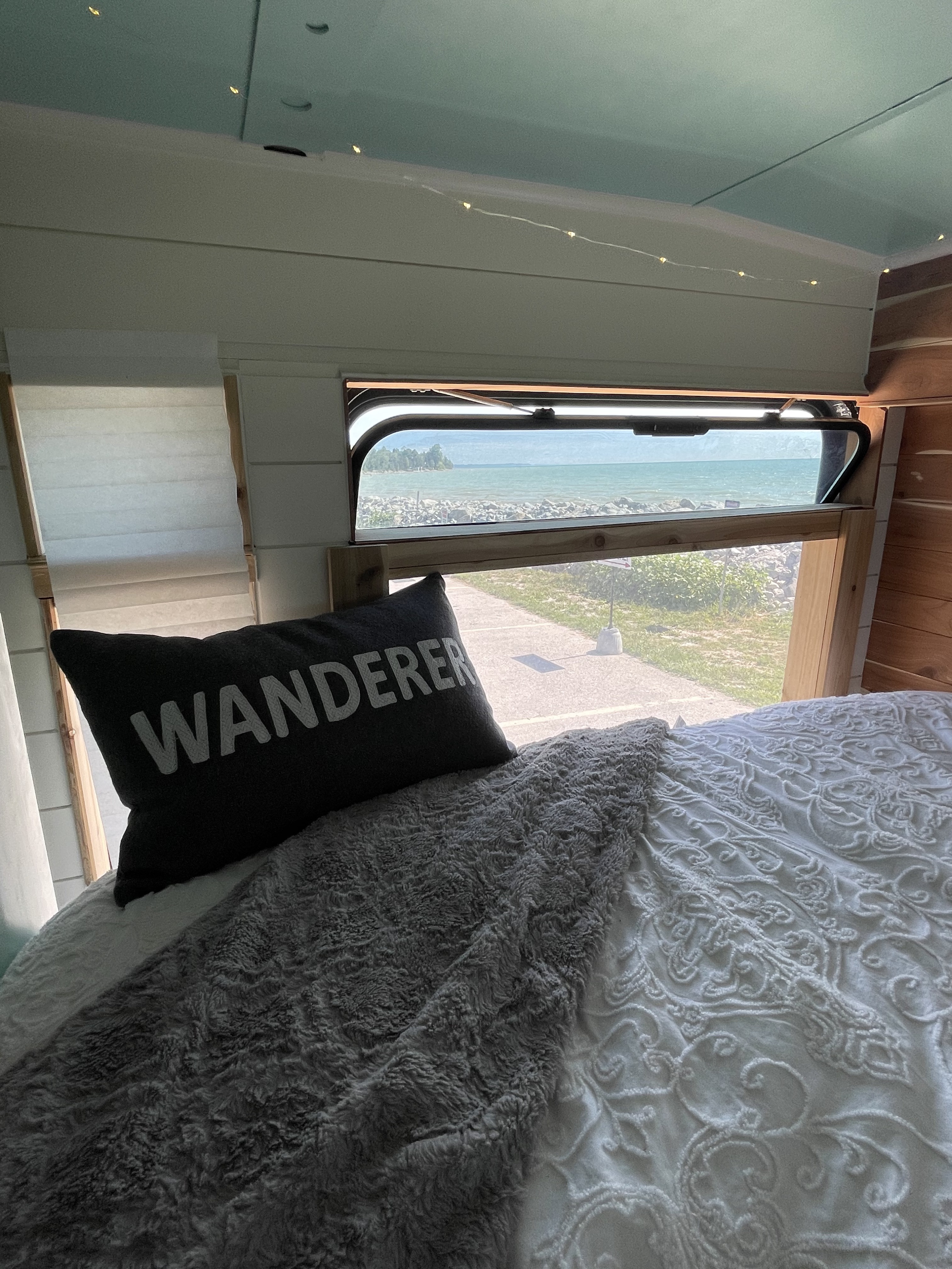 Cozy RV rental bedroom with a 'WANDERER' pillow & fuzzy blanket, offering a stunning water view from the window.. Gillig Low Floor 2021