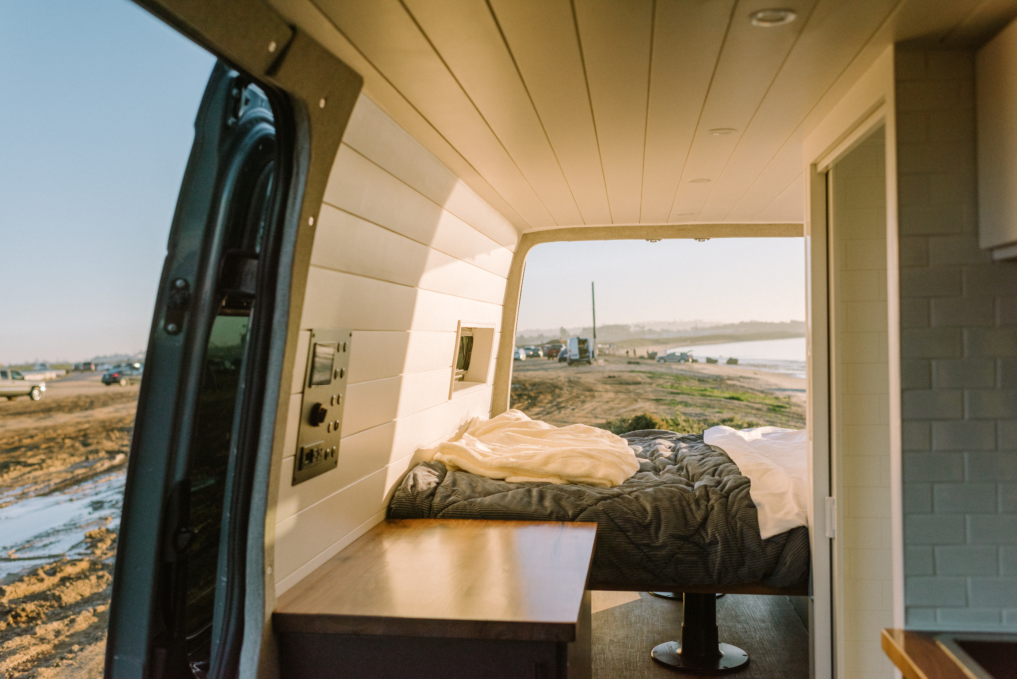 Relax in this RV rental's comfy interior, featuring a bed, wood counter, and open doors to a scenic waterside view.. Mercedes-Benz Sprinter 2021