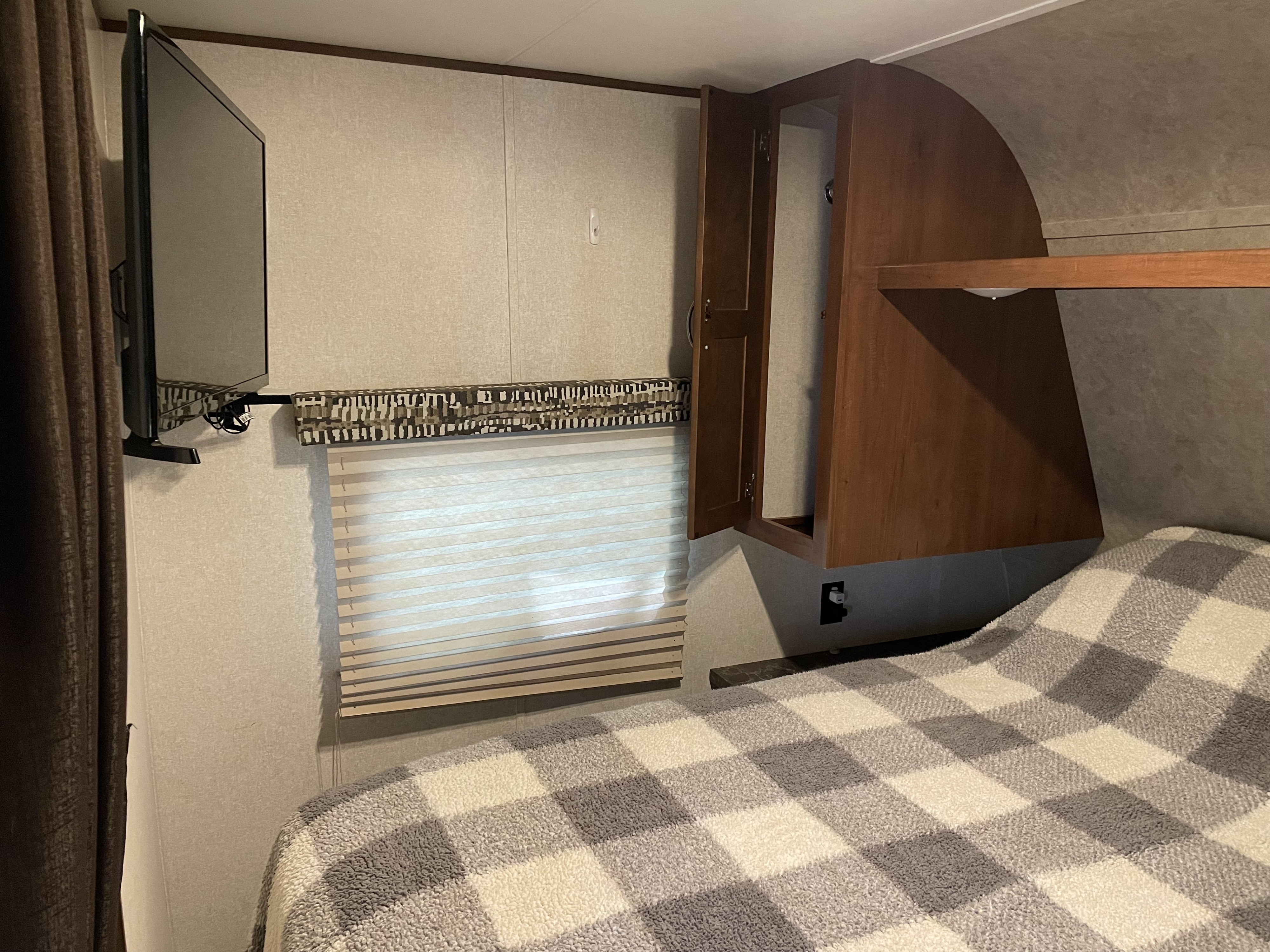 Cozy RV rental bedroom featuring a wall-mounted TV, window with blinds, storage, and a checkered blanket.. Jayco Jay Flight SLX 2017