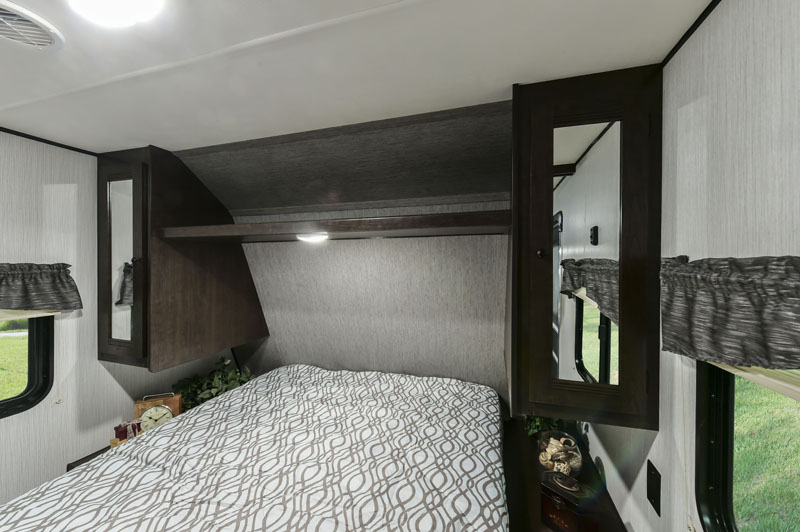 Cozy RV rental bedroom: Features a bed, overhead shelf with light, and side storage cabinets. Windows show green landscape.. Heartland RVs Prowler 2020
