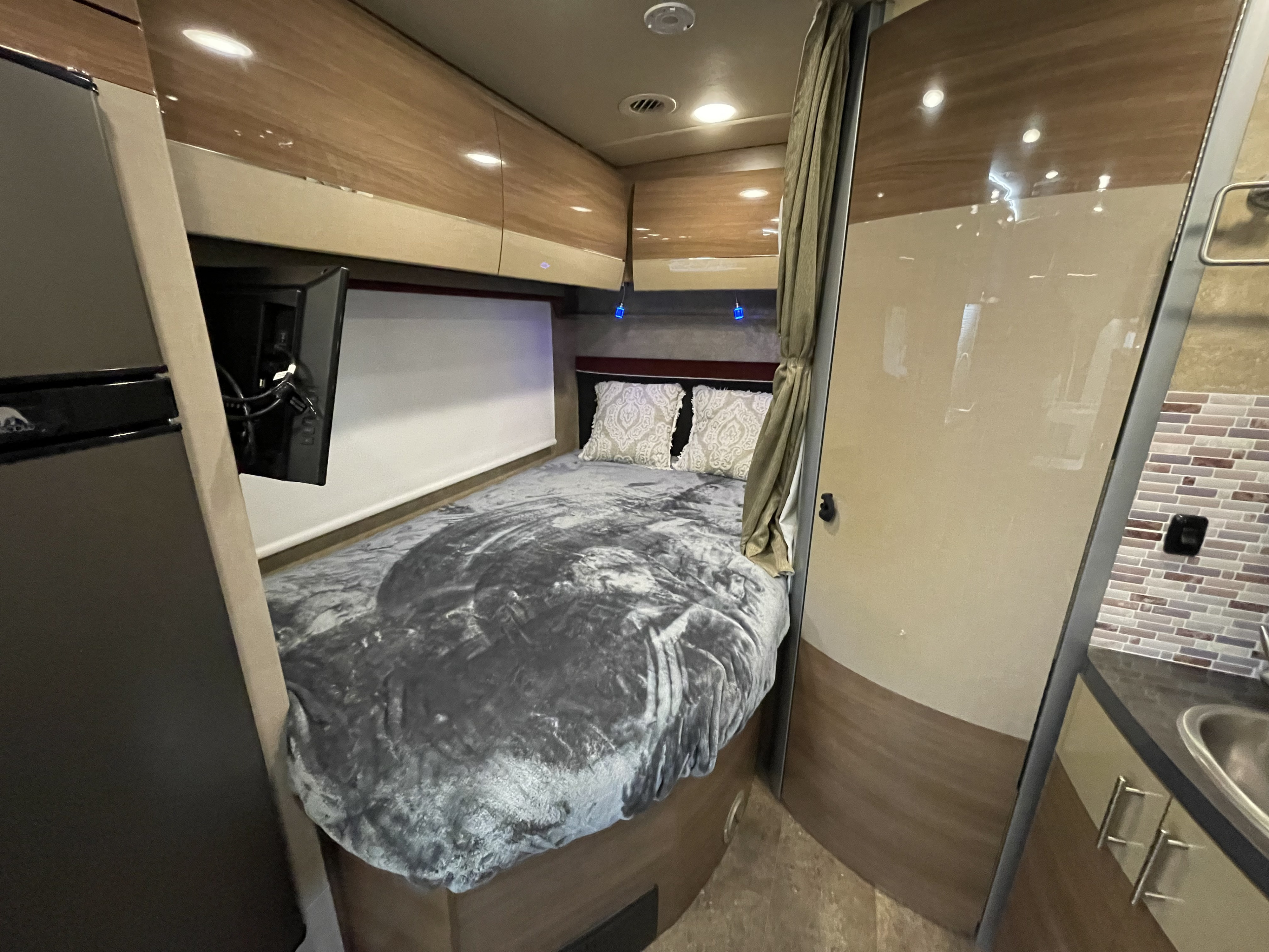 Step inside this cozy rv rental bedroom: a plush bed, wall-mounted TV, overhead storage, and accent lighting for comfortable travel.. Itasca Navion 2016
