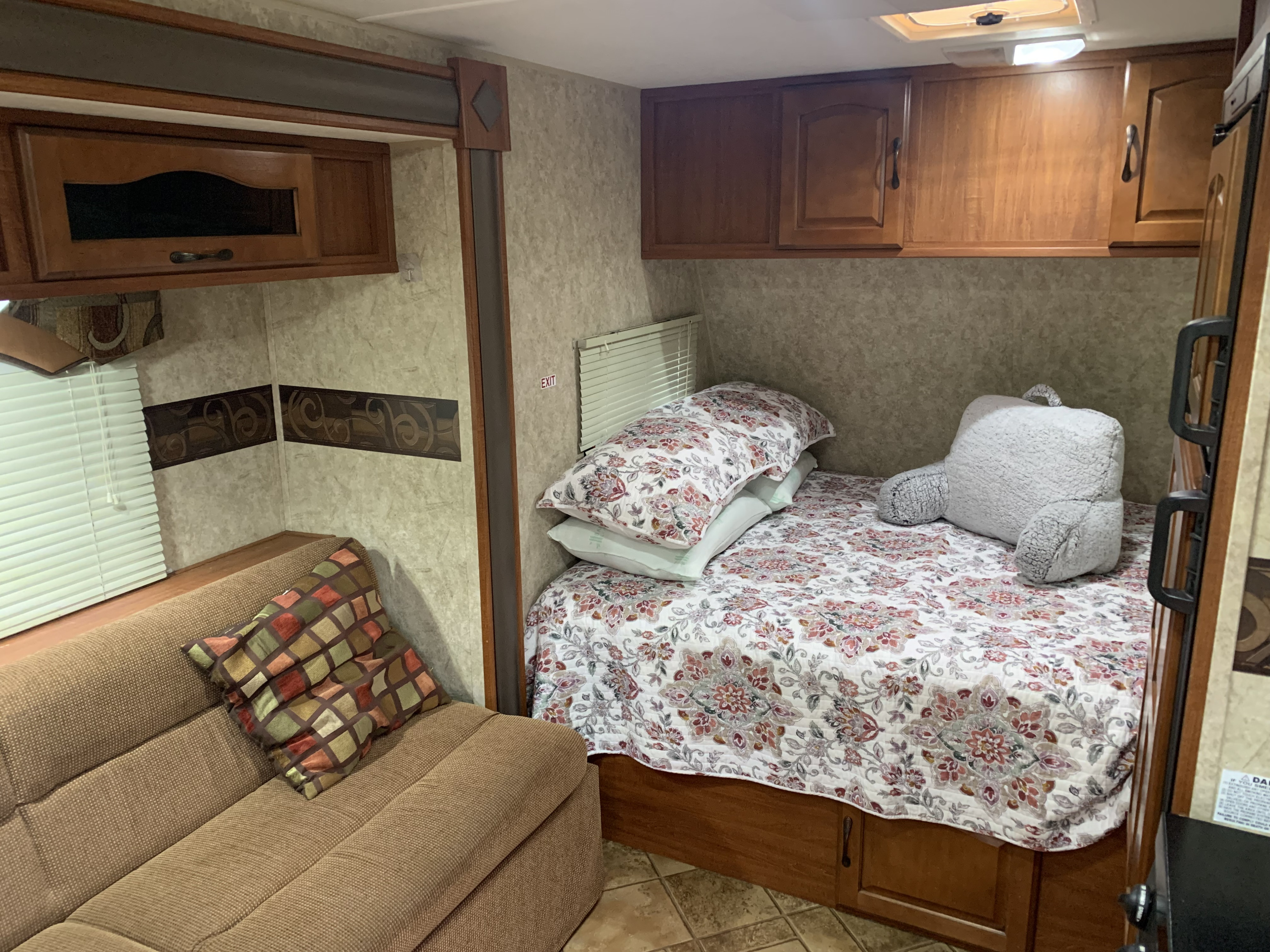 Cozy RV rental interior features a bed with floral comforter, gray back pillow, a sofa, and wood cabinets. Tiled floor.. Forest River Surveyor 2015