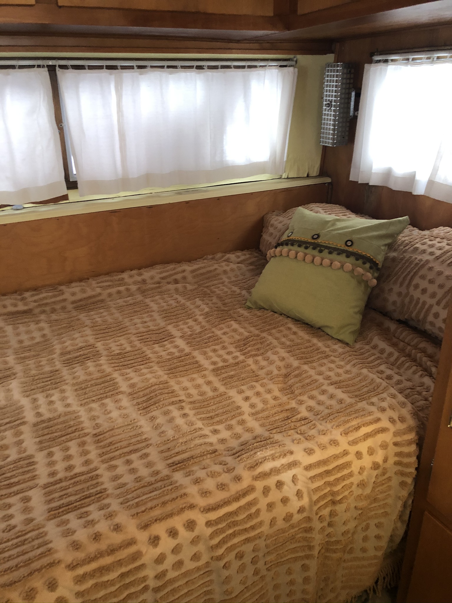 Inside a cozy RV rental: a bed with a textured brown blanket & green pillow. Bright windows with white curtains.. Holiday Rambler Traveler 1964