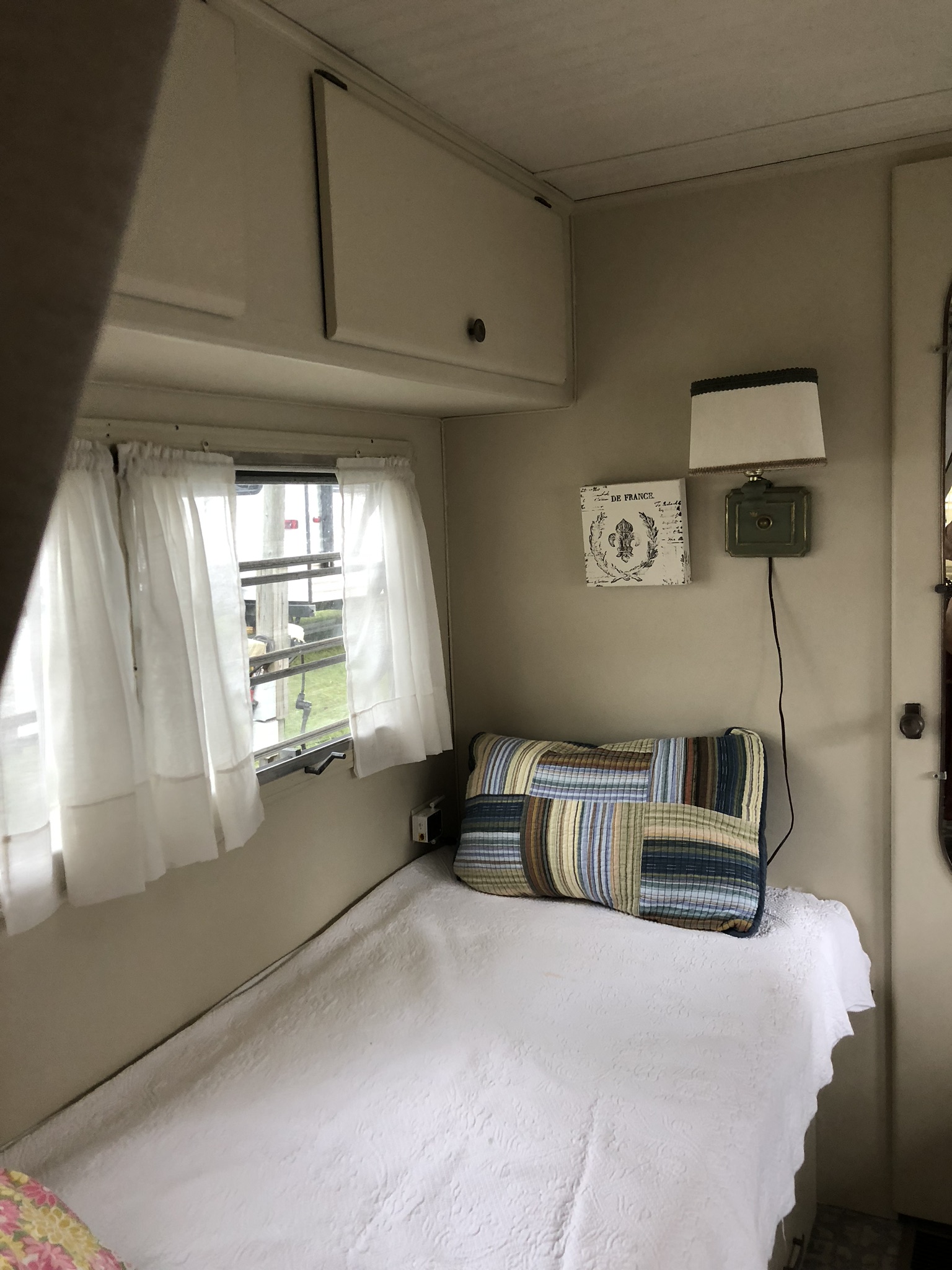 This RV rental interior features a cozy single bed, wall lamp, and window with curtains, offering a compact stay.. Holiday Rambler Vacationer 1969
