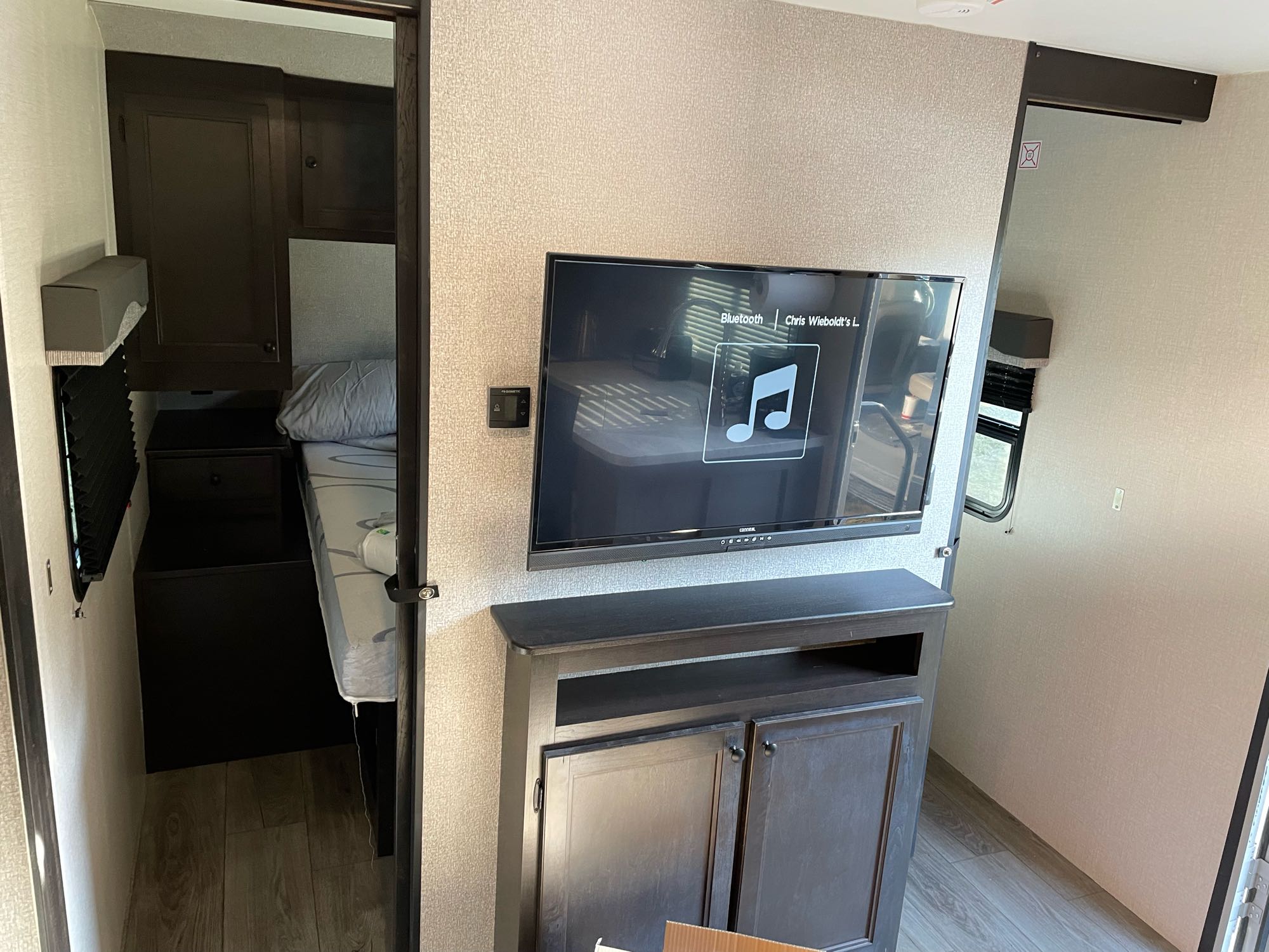 View inside an RV rental: a bedroom with a bed is seen through an open doorway, beside a wall-mounted TV and storage cabinet.. Highland Ridge Mesa Ridge 2021