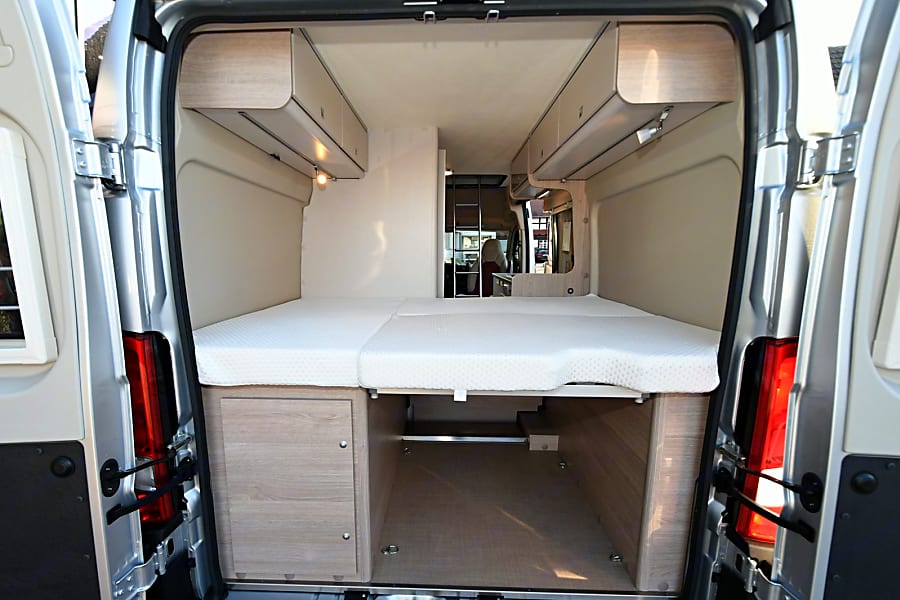 View into a modern RV rental camper van showing a comfortable bed, overhead storage, and accessible under-bed space.. Bürstner Campeo 600 2024