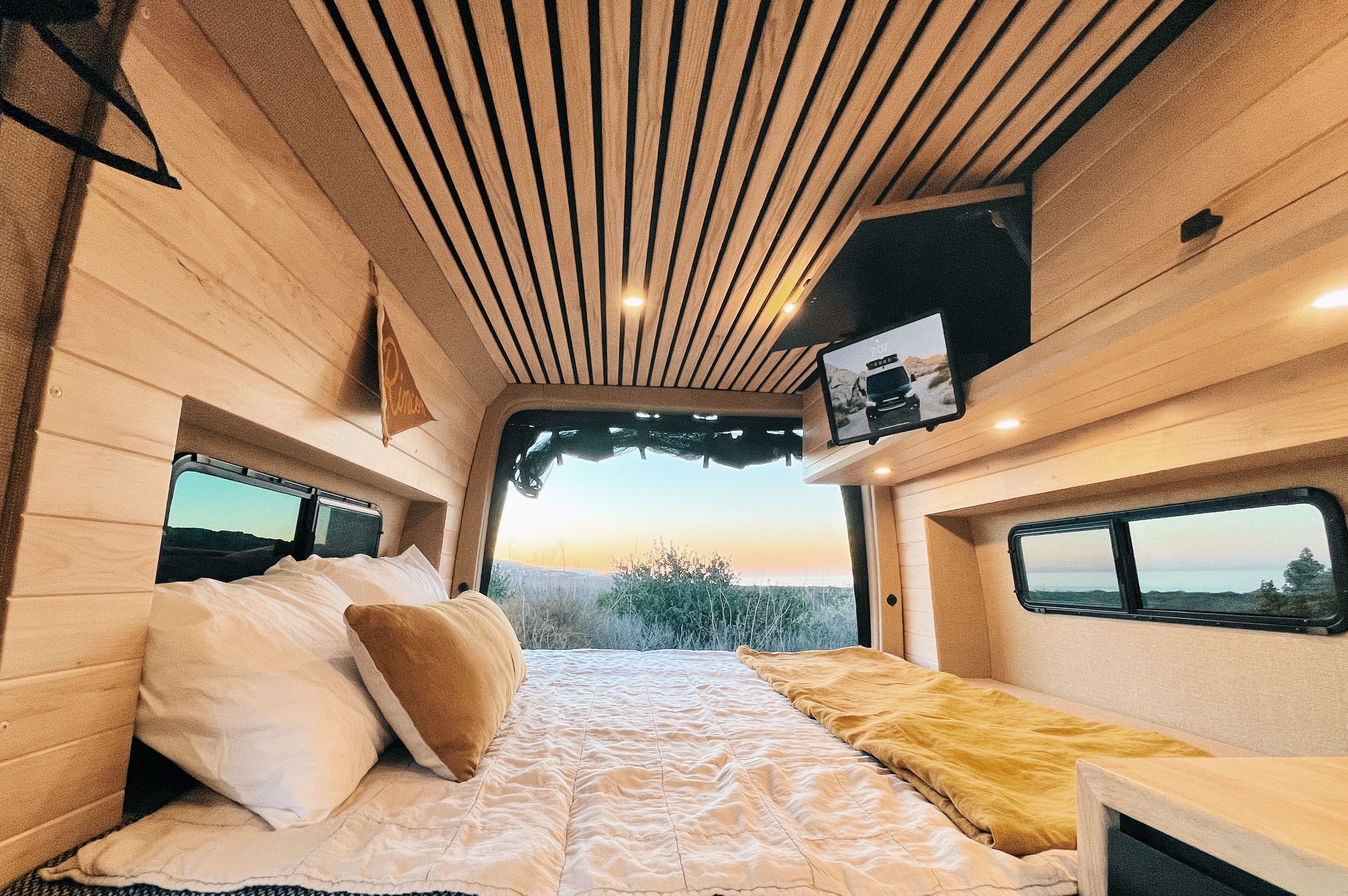 Cozy RV rental interior: wood-paneled walls, comfy bed, and panoramic window view of a serene sunset landscape.. Mercedes-Benz Sprinter 2021