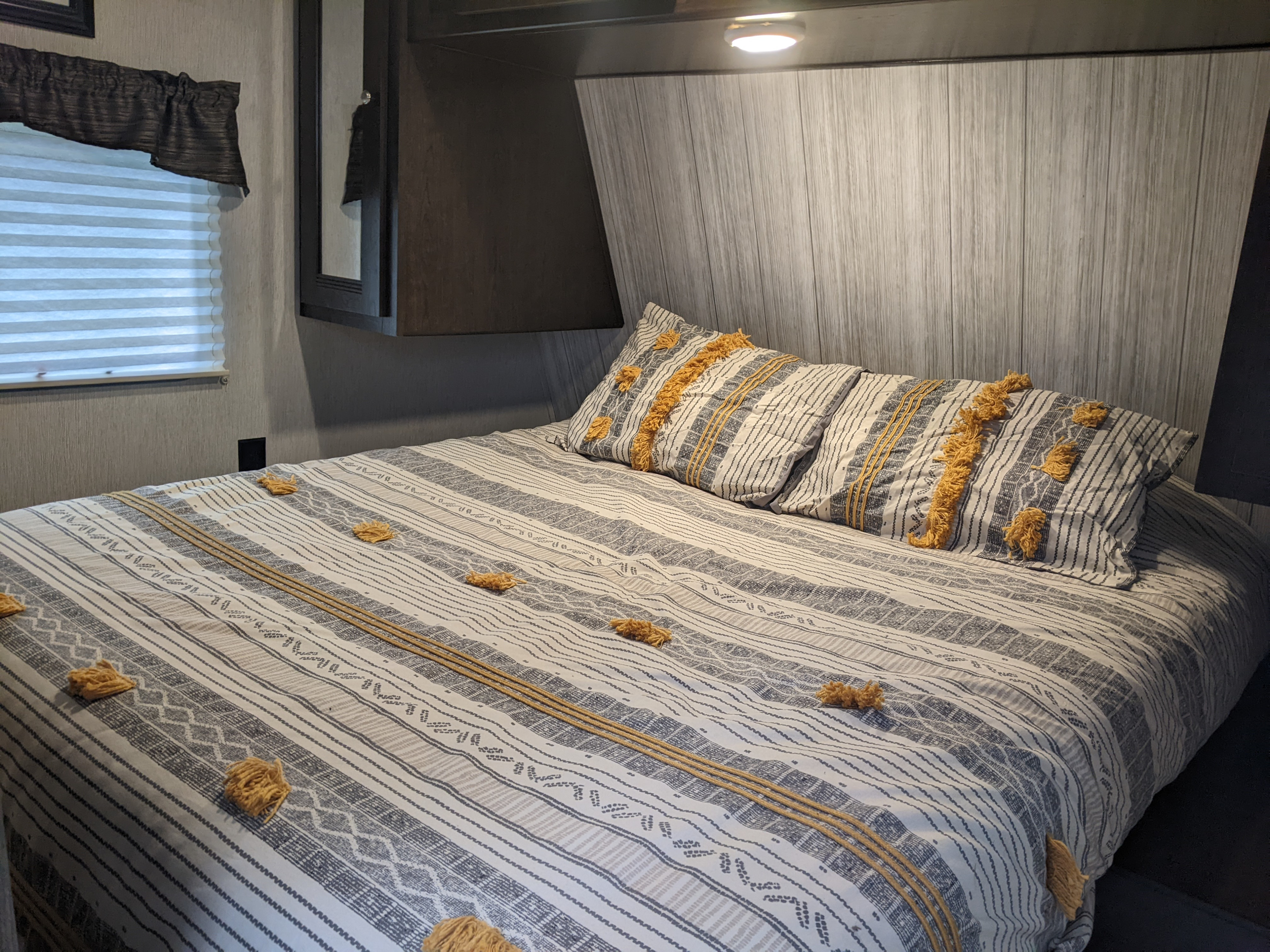 Cozy RV rental bedroom: bed with striped and tufted bedding, overhead cabinets, and a window with blinds.. Heartland RVs Pioneer 2019