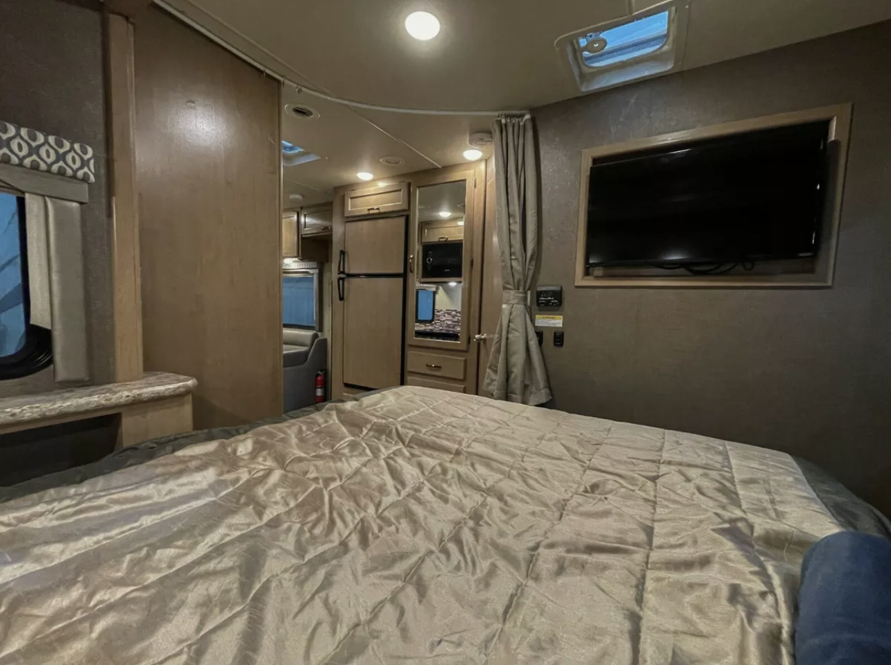 Cozy RV rental interior showing a bedroom with a made bed and wall-mounted TV, looking towards the kitchenette and living area.. Thor Motor Coach Freedom Elite 2019