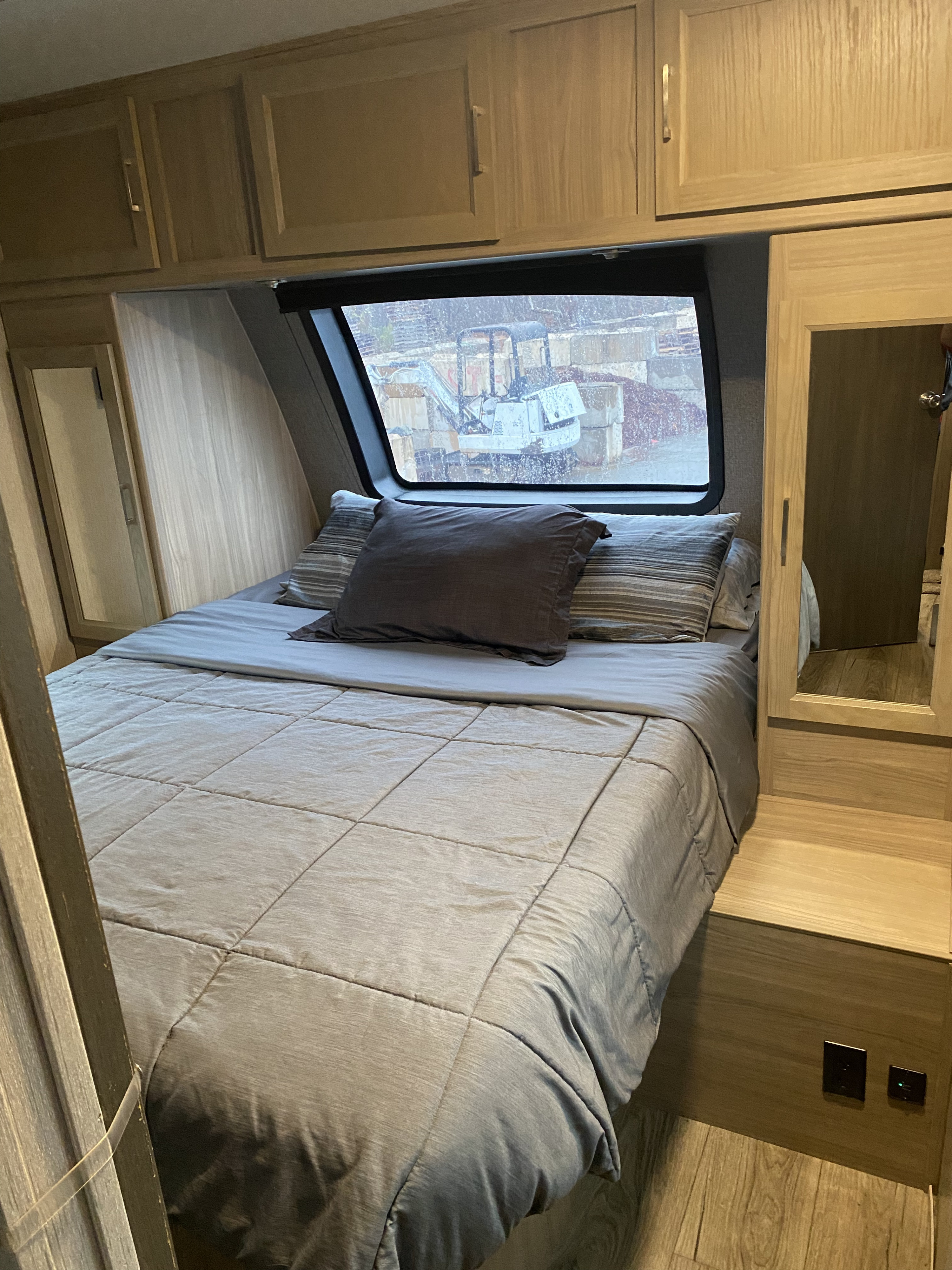 An RV rental bedroom: bed with grey comforter & pillows, wooden cabinets, mirrored closet. Window view of machinery.. Coachmen Liberty 2020