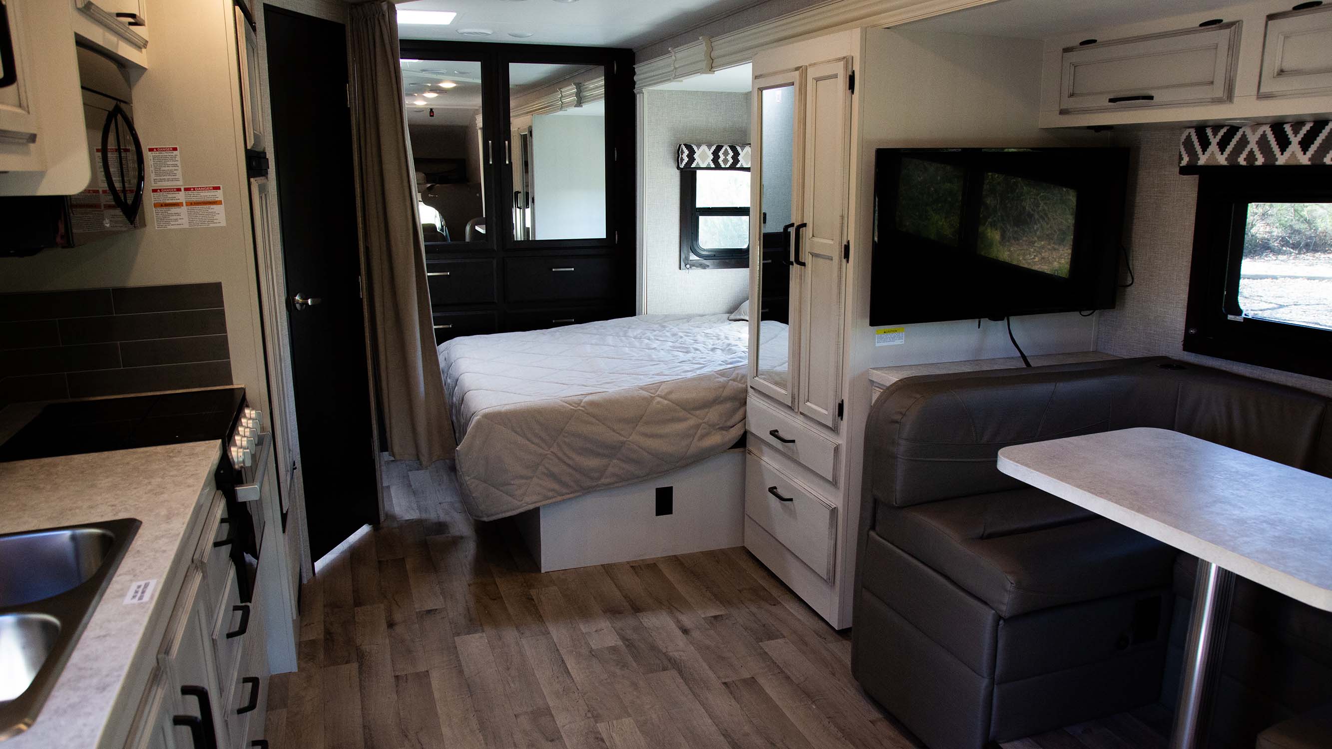 Inside a modern RV rental: view of the compact kitchen, cozy bed, and dining booth with TV.. Jayco Redhawk 2022