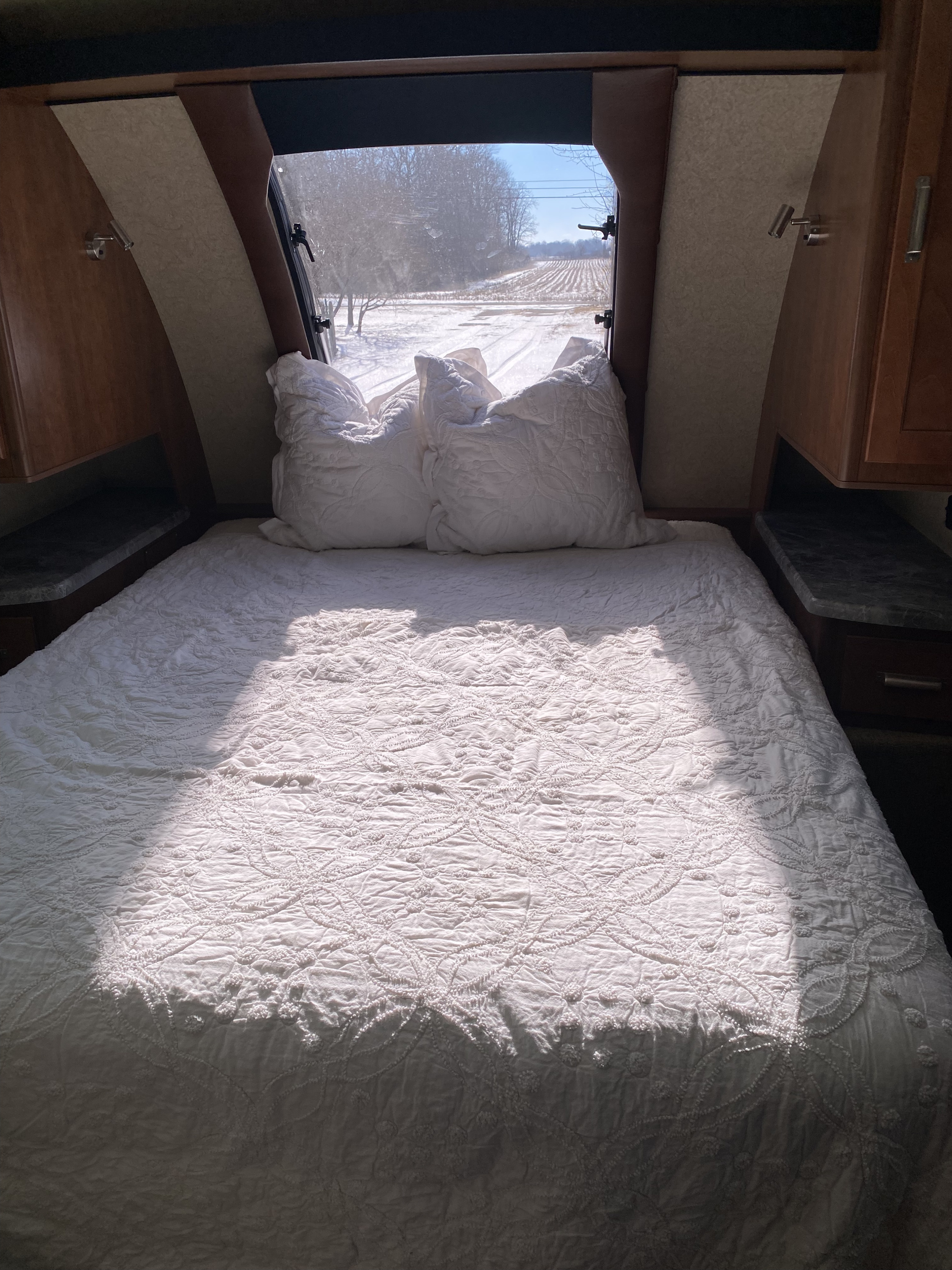 Cozy RV rental bedroom featuring white bedding, reading lights, and a serene snowy landscape view from the back window.. Lance Manufacturing 2375 2021