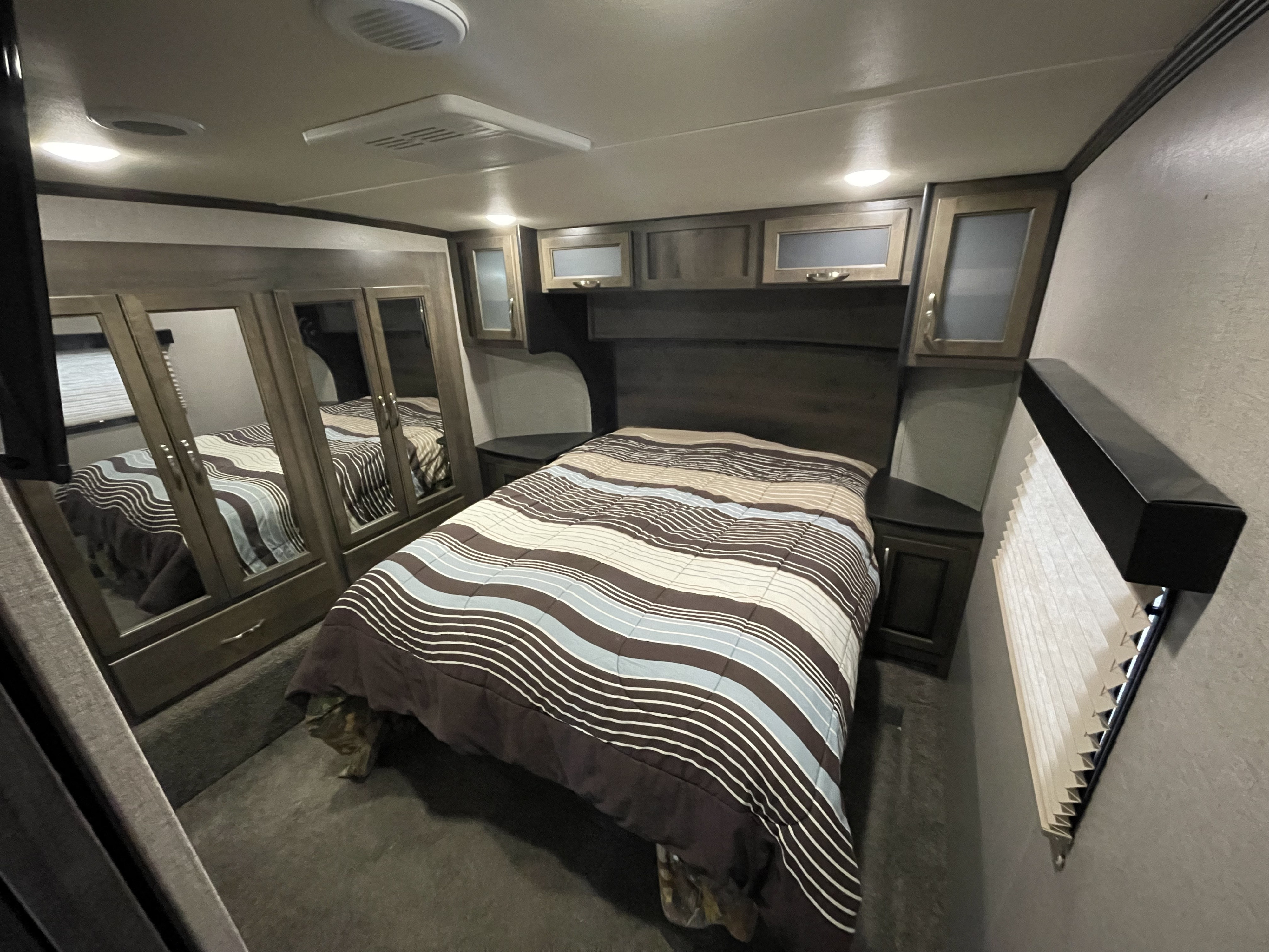 Cozy RV rental bedroom with striped bed, overhead cabinets, and mirrored wardrobe. Comfortable space inside a camper.. Dutchmen Voltage 2018