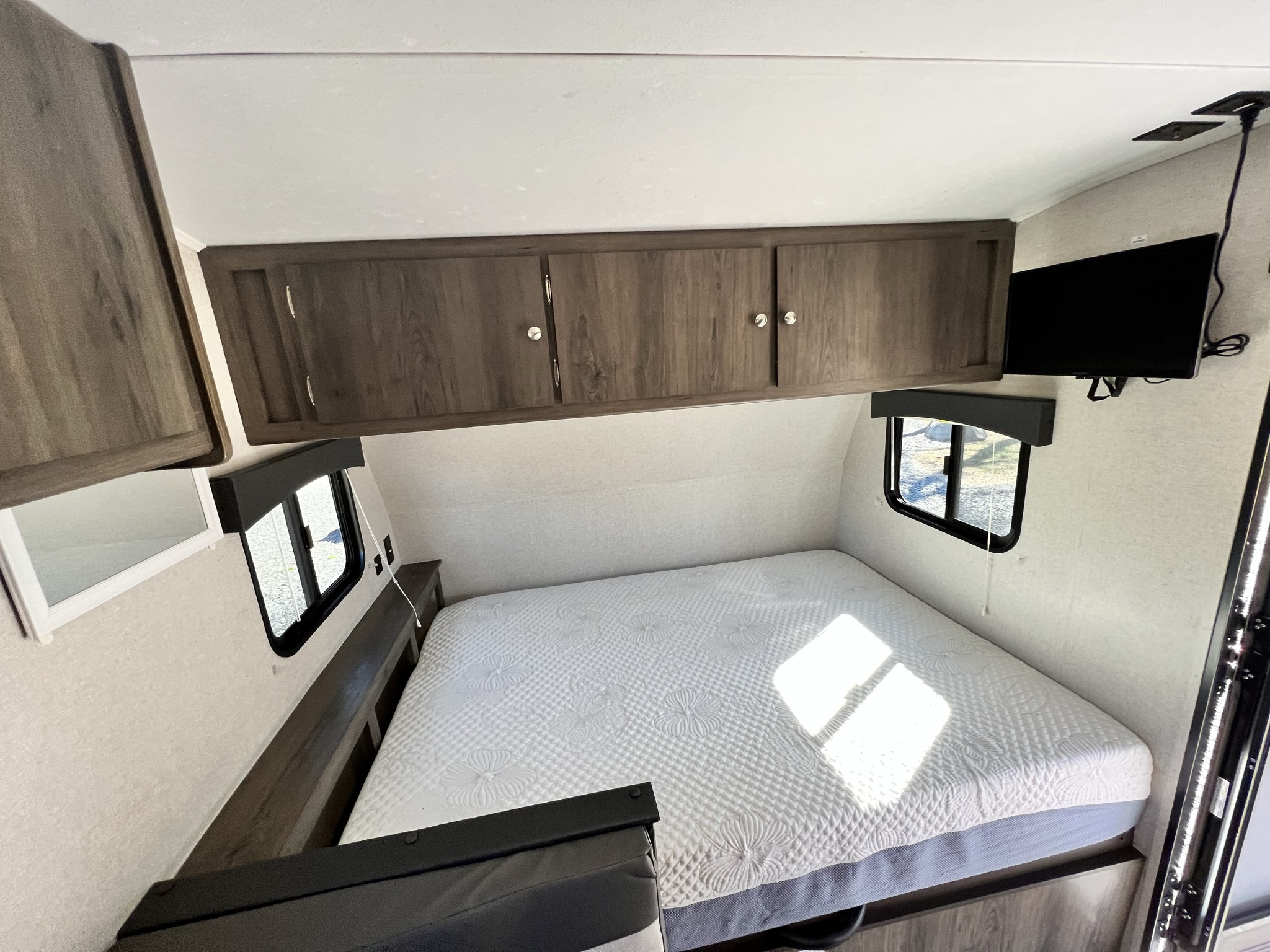 Step into this RV rental's bedroom: a bright space with a quilted queen bed, wall-mounted TV, and overhead cabinets.. Forest River 17BH 2021
