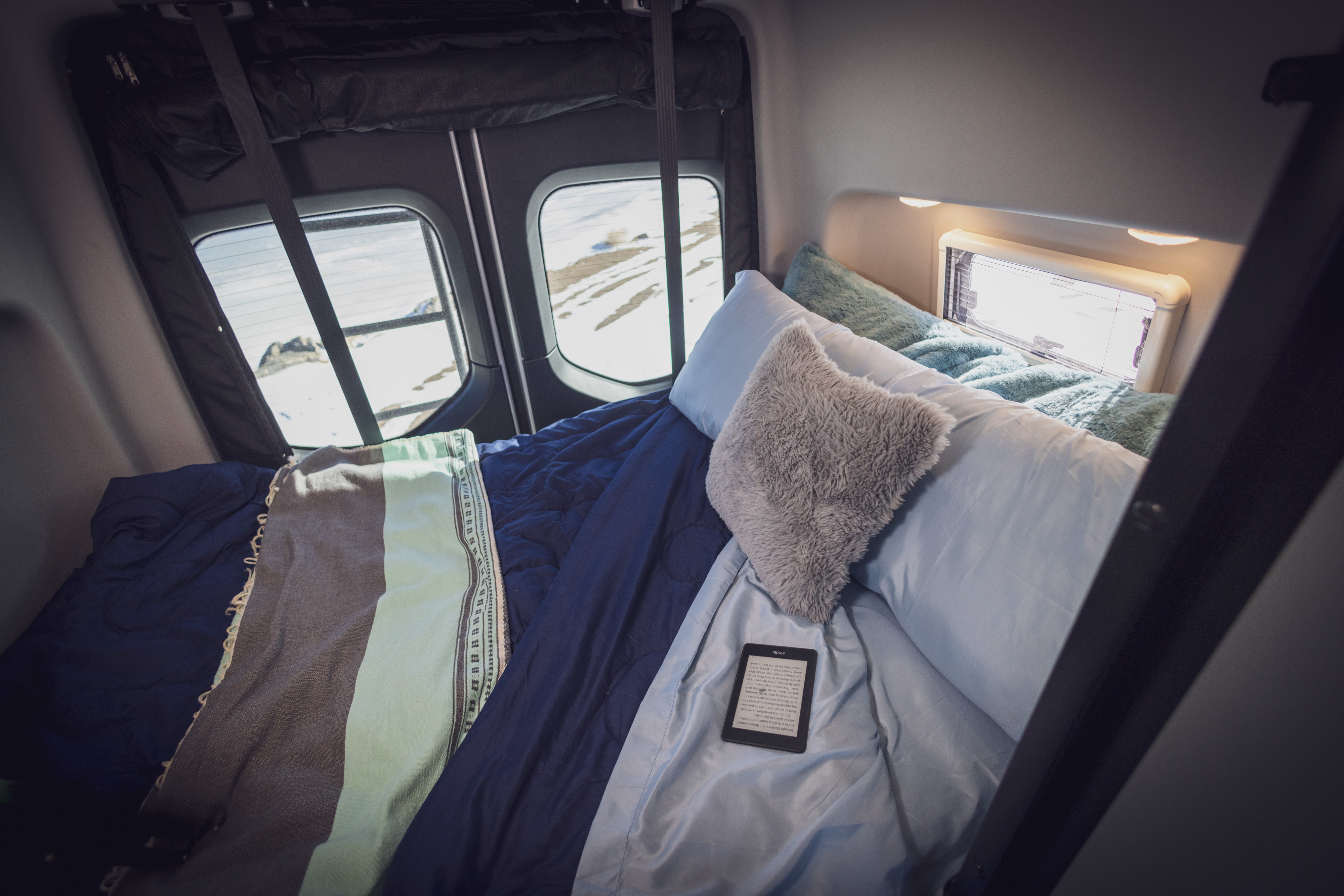 Cozy RV rental bed with a view of a snowy landscape and an e-reader, perfect for a winter escape in a camper van.. Winnebago Revel 2022