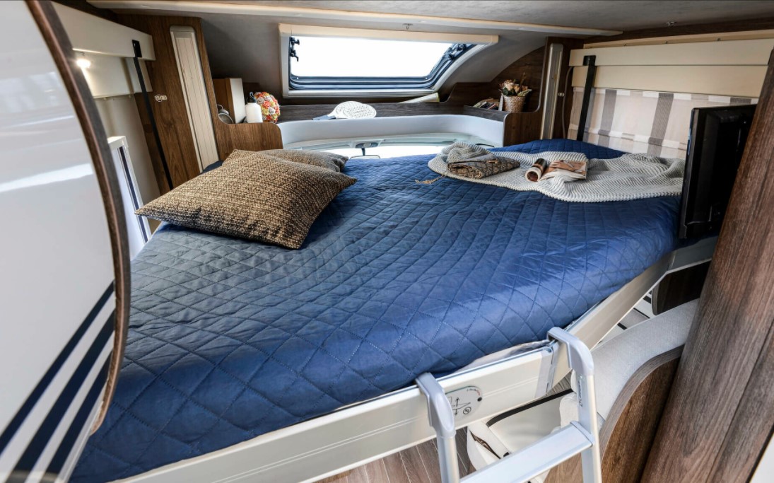 Inside an RV rental: a cozy, elevated bed with blue quilt, pillows, window, and TV, offering a comfortable sleeping space for travel.. Roller Team T-line 700 2025