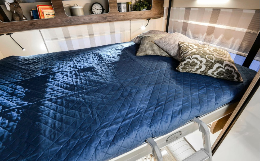 Relax in this RV rental's comfy bunk bed, featuring a quilted blue spread, pillows, and overhead shelving.. Roller Team T-line 700 2025