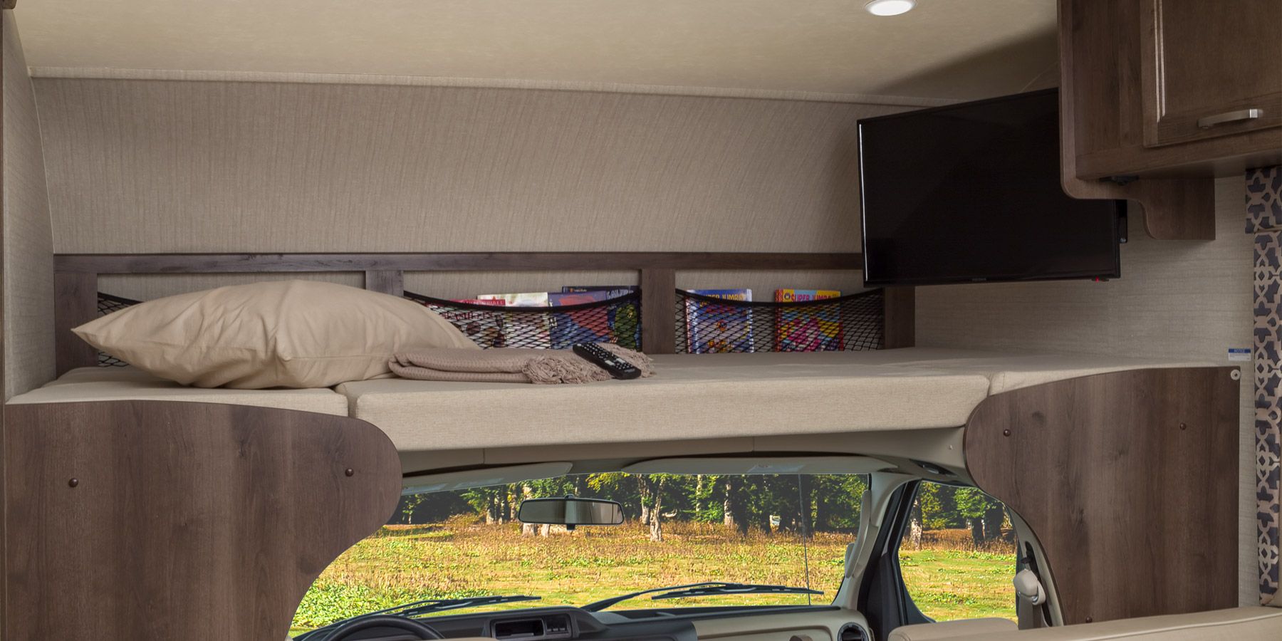 Inside an RV rental: a cozy cab-over bunk with bed, pillow, TV, and handy mesh storage for books, overlooking a scenic view.. Jayco Redhawk 2018