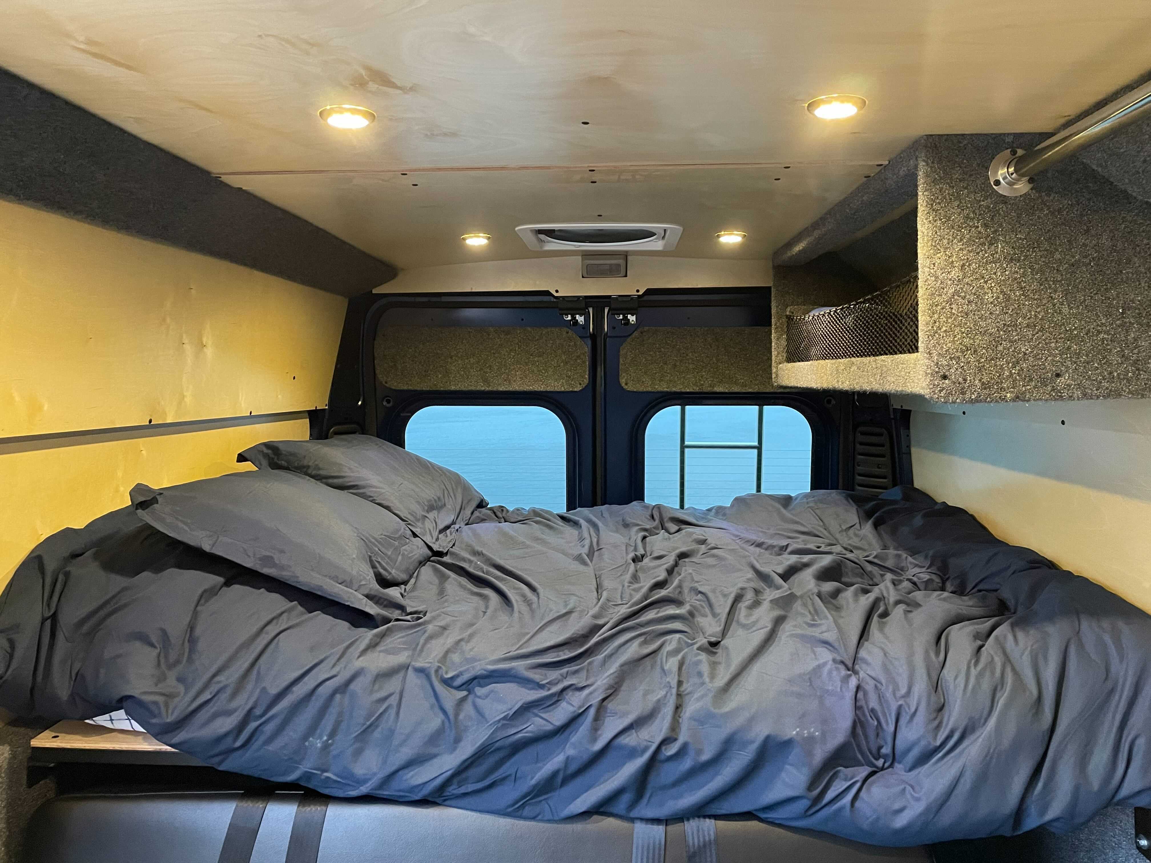 Step into comfort! This RV rental features a cozy bed with dark bedding, custom wood paneling, and practical overhead storage.. Dodge Ram Promaster 250020 2021