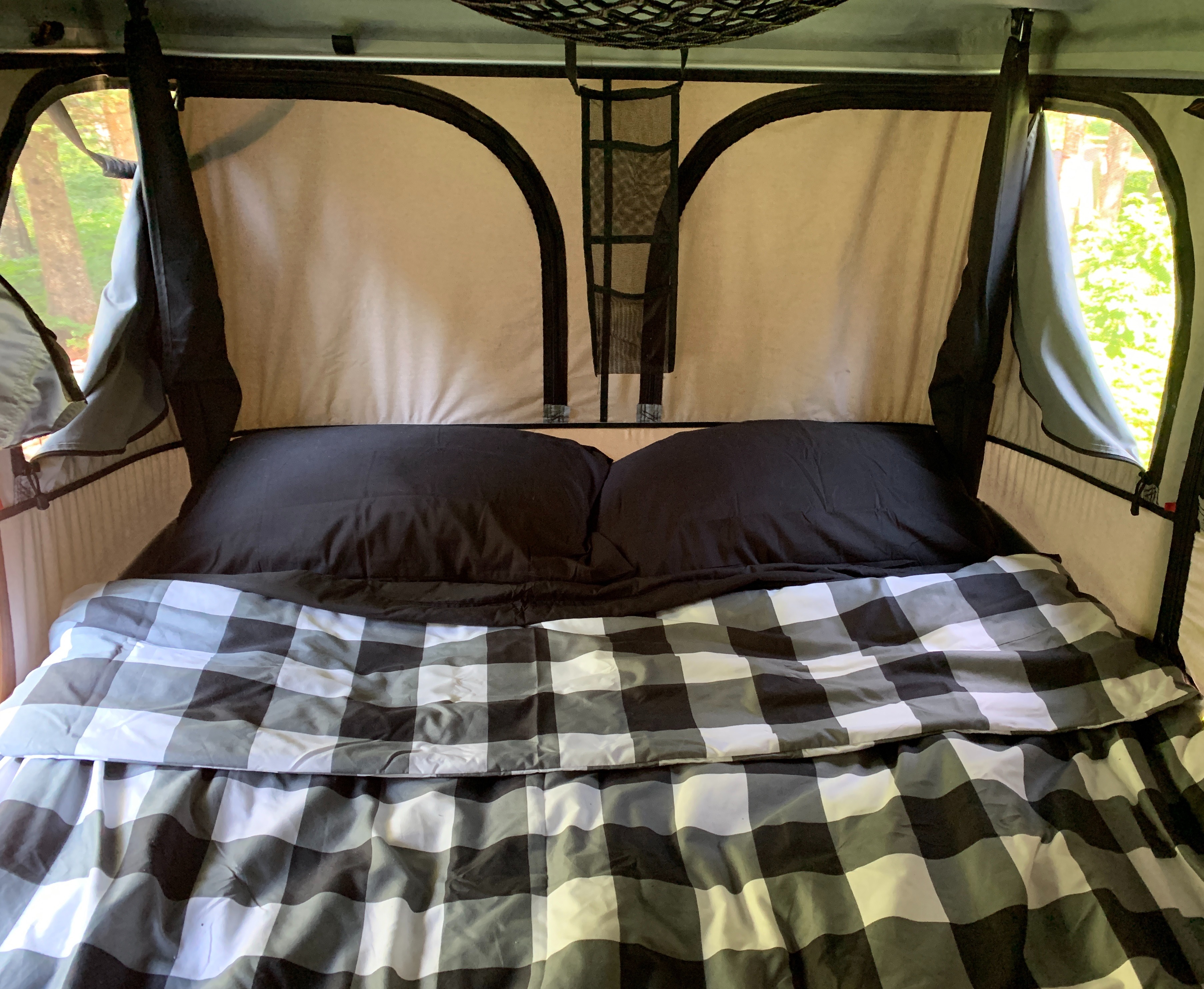 Cozy RV rental interior with a comfortable bed, black pillows, and a buffalo check comforter. Windows reveal green trees.. Dodge Grand Caravan 2018