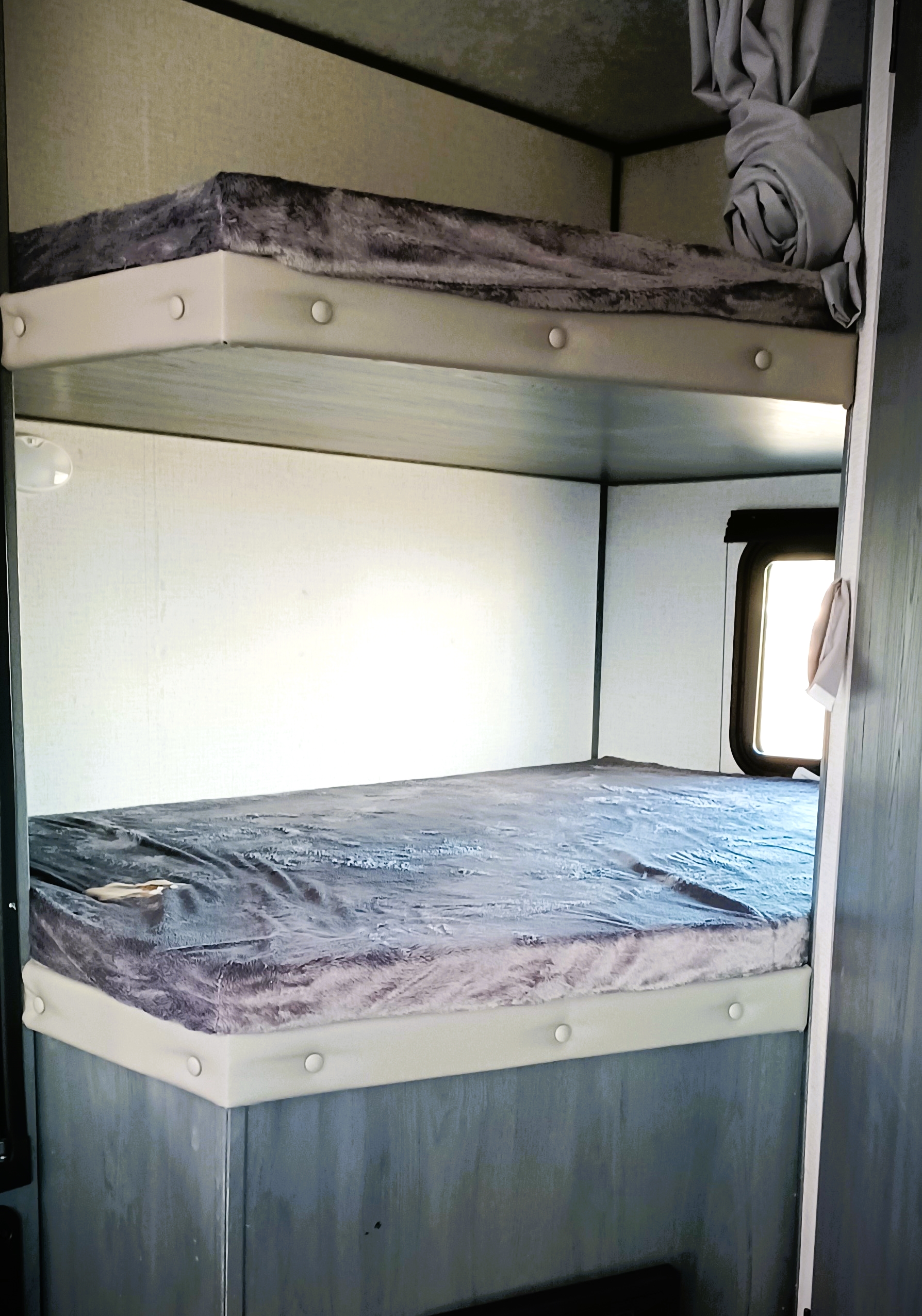View of two stacked bunk beds inside an RV rental. Each bunk features a grey furry cover, with light walls and a window visible.. Dutchmen Aspen Trail 2021