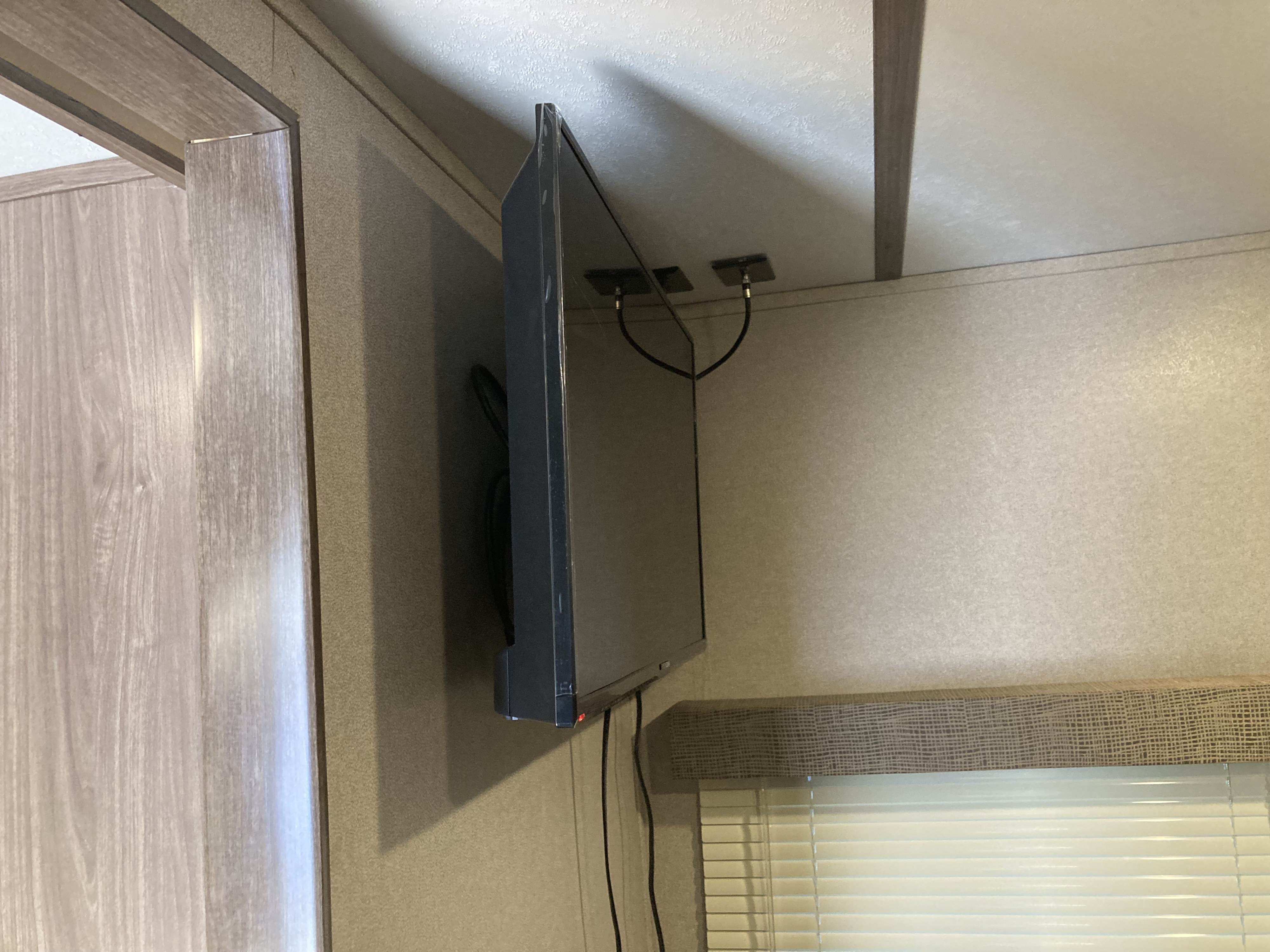 Wall-mounted TV in an RV rental, with visible cables connected overhead. A wood-like frame and window blinds are also visible.. Keystone Hideout 2019