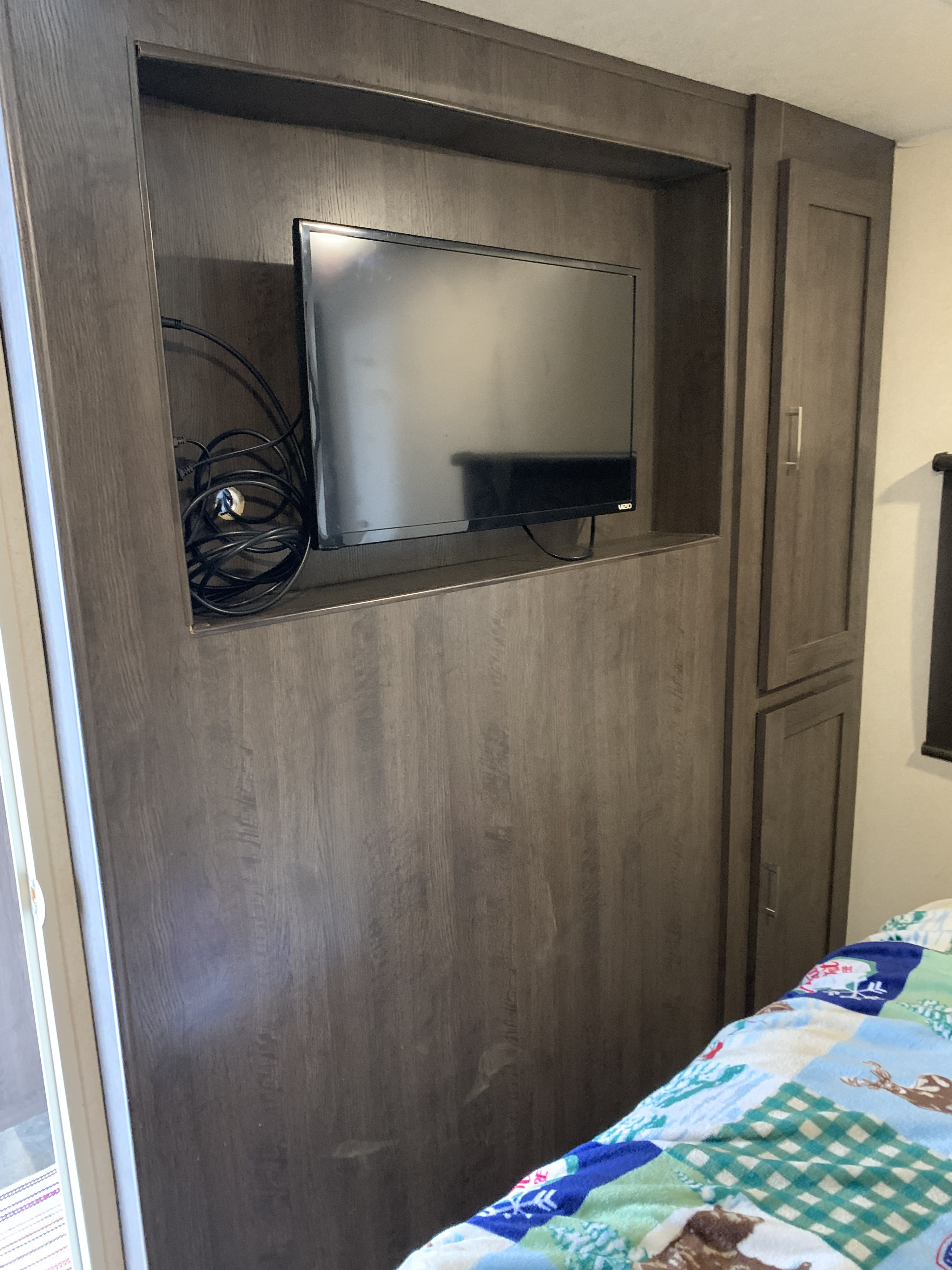 RV rental bedroom: wood-paneled wall unit with TV, tangled cables, storage cabinets, and a colorful bedspread.. Forest River Wildwood 2018