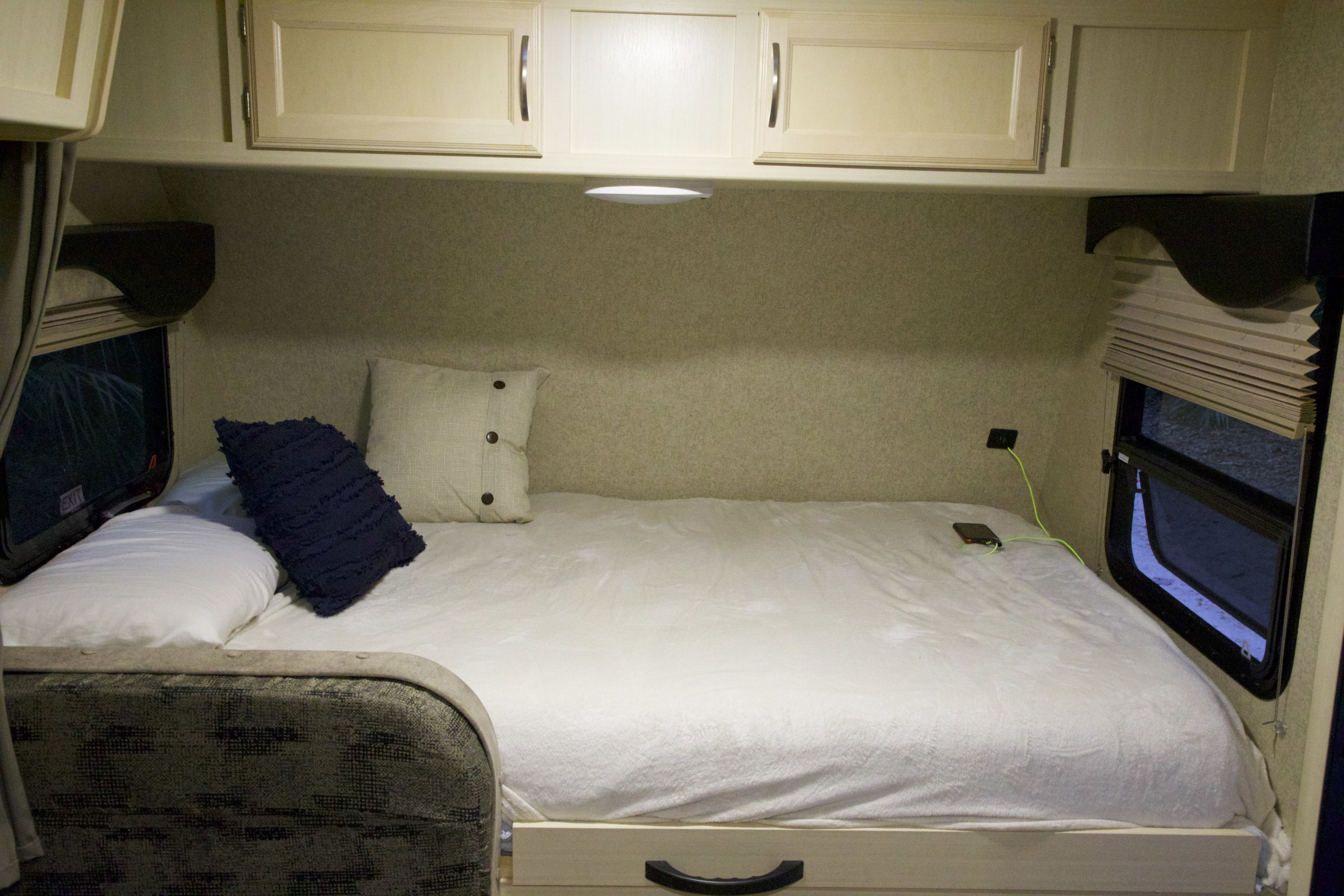 Cozy RV rental bedroom showing a bed with white linens, pillows, overhead cabinets, and windows. A phone charges on the right.. Winnebago Micro Minnie 2018