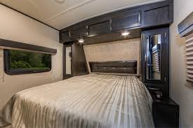 Modern RV rental bedroom with a striped queen bed, overhead cabinets, side mirrors, and a window, offering comfort for travel.. Grand Design Imagine 2800BH 2019