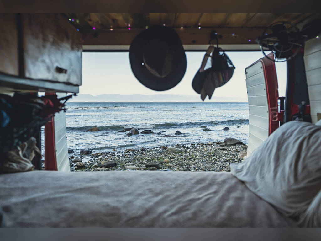 View from inside an RV rental shows a bed in foreground, beyond it a rocky beach, ocean, and distant mountains.. Dodge promaster 3500 2018