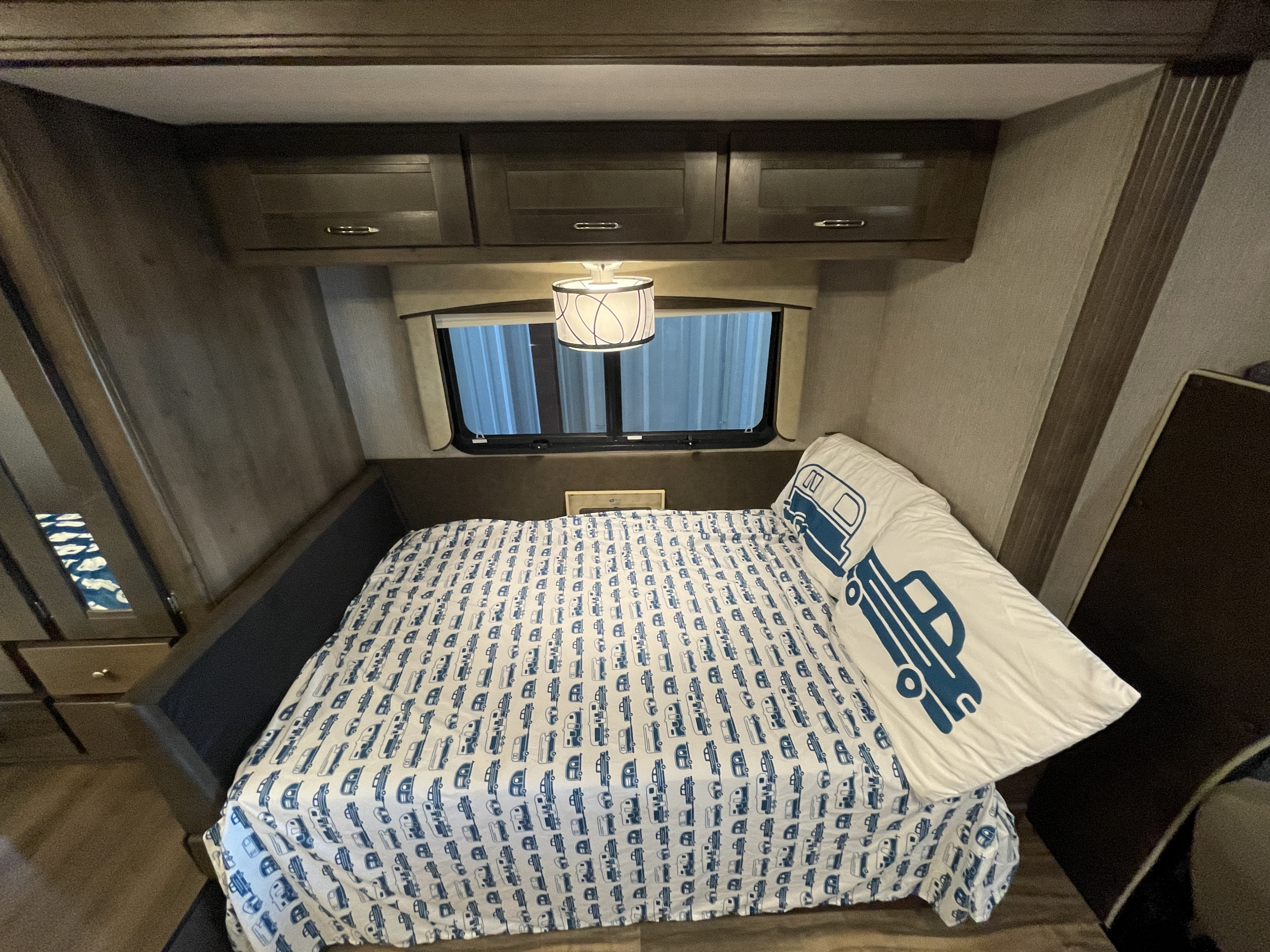 Cozy RV rental bedroom featuring camper-themed bedding, overhead cabinets, and a window for comfortable travel.. Nexus Triumph 2021