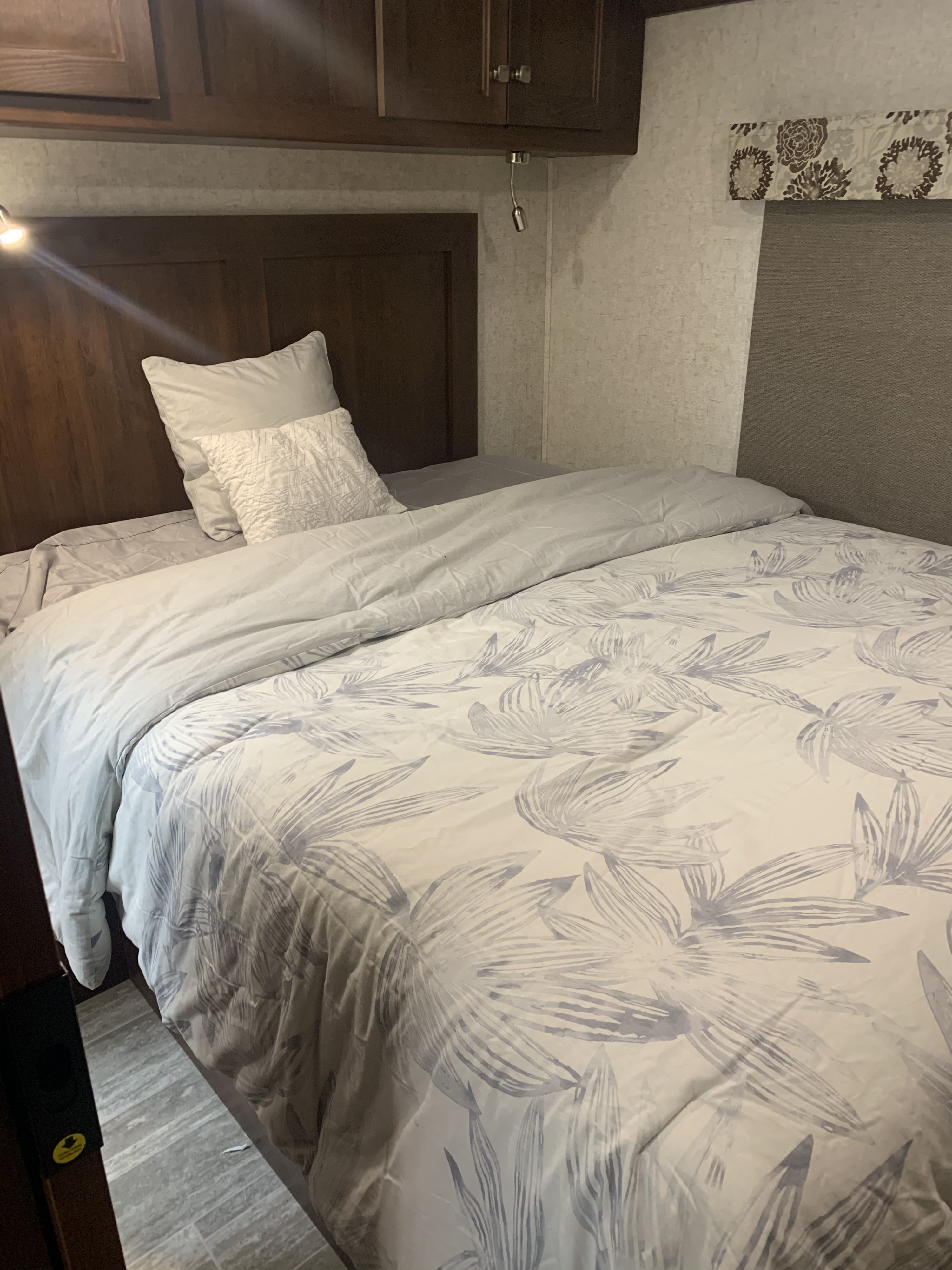 A neatly made bed inside an RV rental, featuring light grey leaf-patterned bedding, pillows, and dark wood cabinetry.. Forest River Forester 2019
