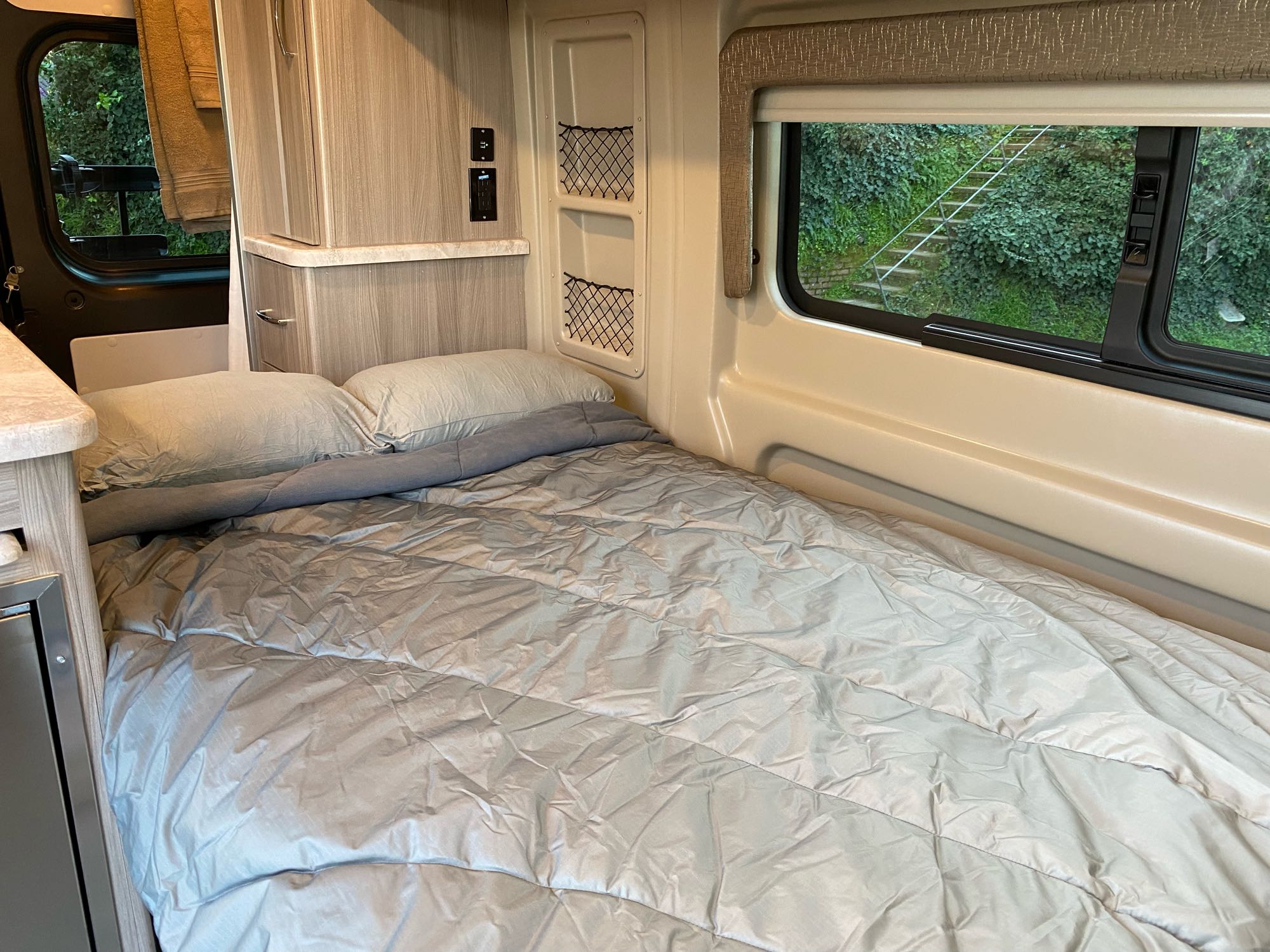 This RV rental interior features a comfortable grey bed, cabinetry, and a window showing a green, ivy-covered outdoor view.. Thor Motor Coach Scope 2022
