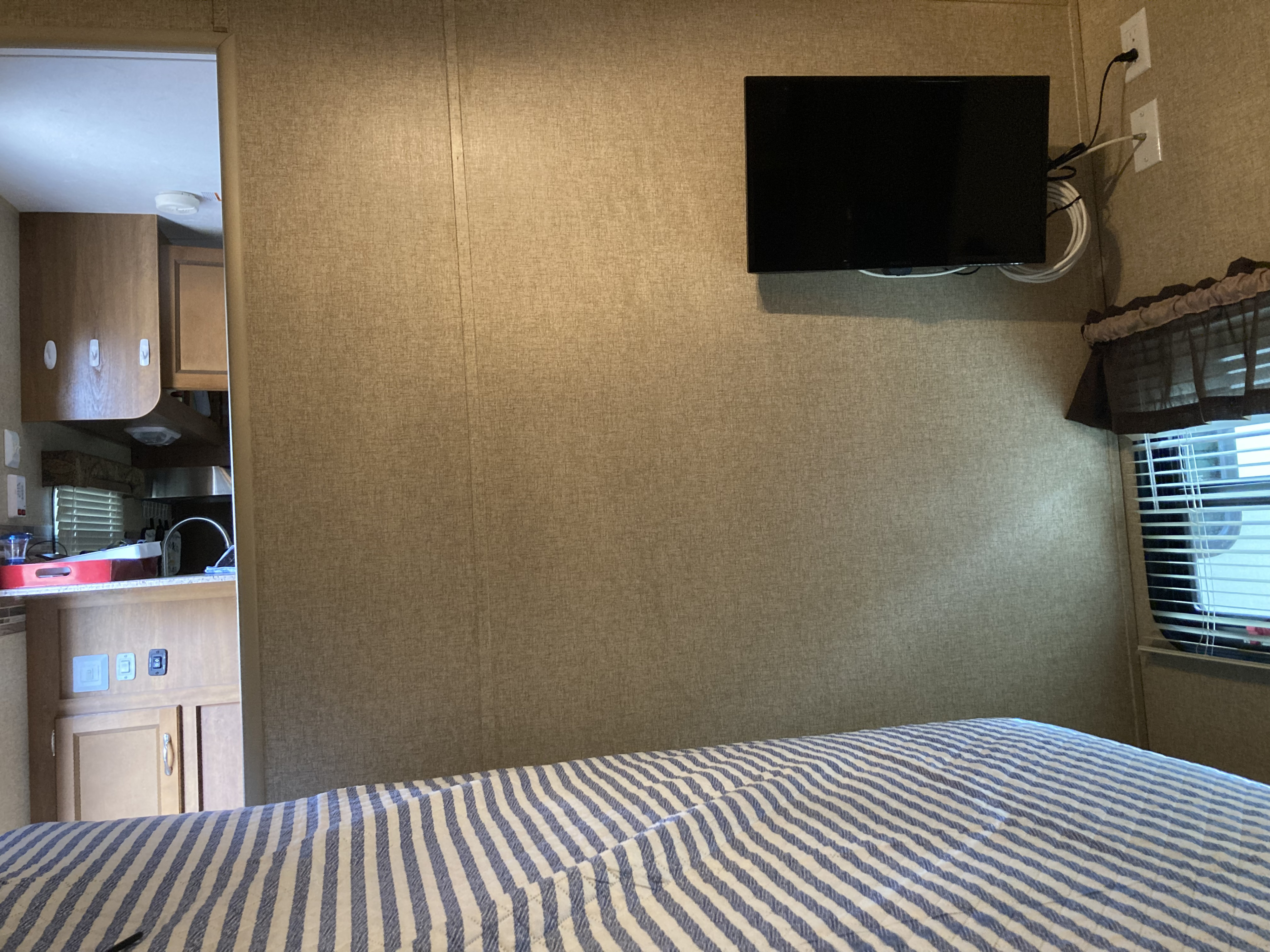 RV rental interior showcasing a cozy bedroom with striped bedding, wall-mounted TV, and adjacent compact kitchen/living area.. Coachmen Catalina 2015