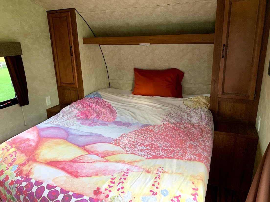 A vibrant RV rental bedroom with a colorful artistic duvet, built-in wooden cabinets, and a side window view.. Gulf Stream Amerilite 2015