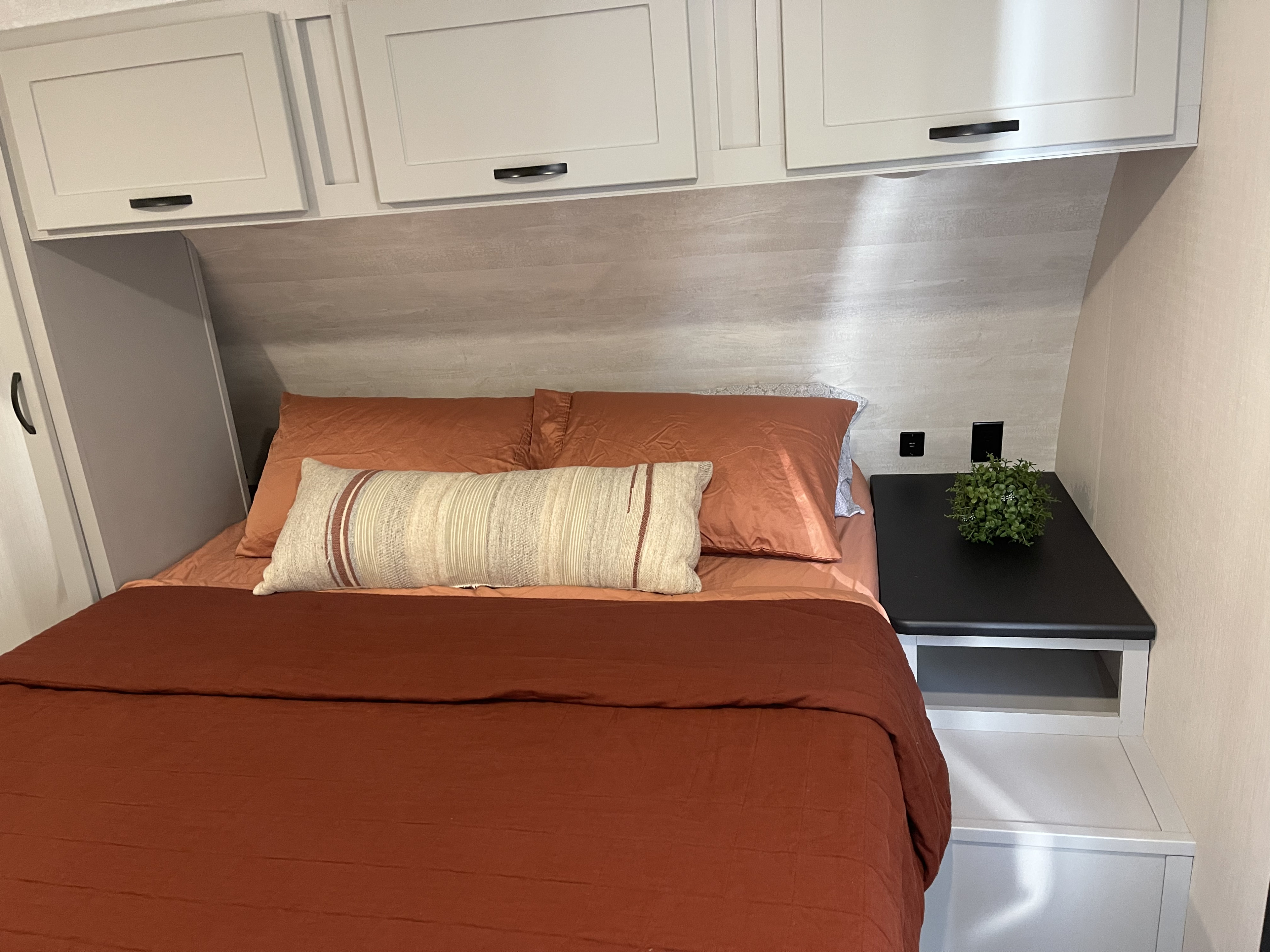 Cozy RV rental bedroom features a bed with rust-colored bedding, modern white cabinets, and a neat nightstand.. Forest River Vibe Travel Trailer 2022