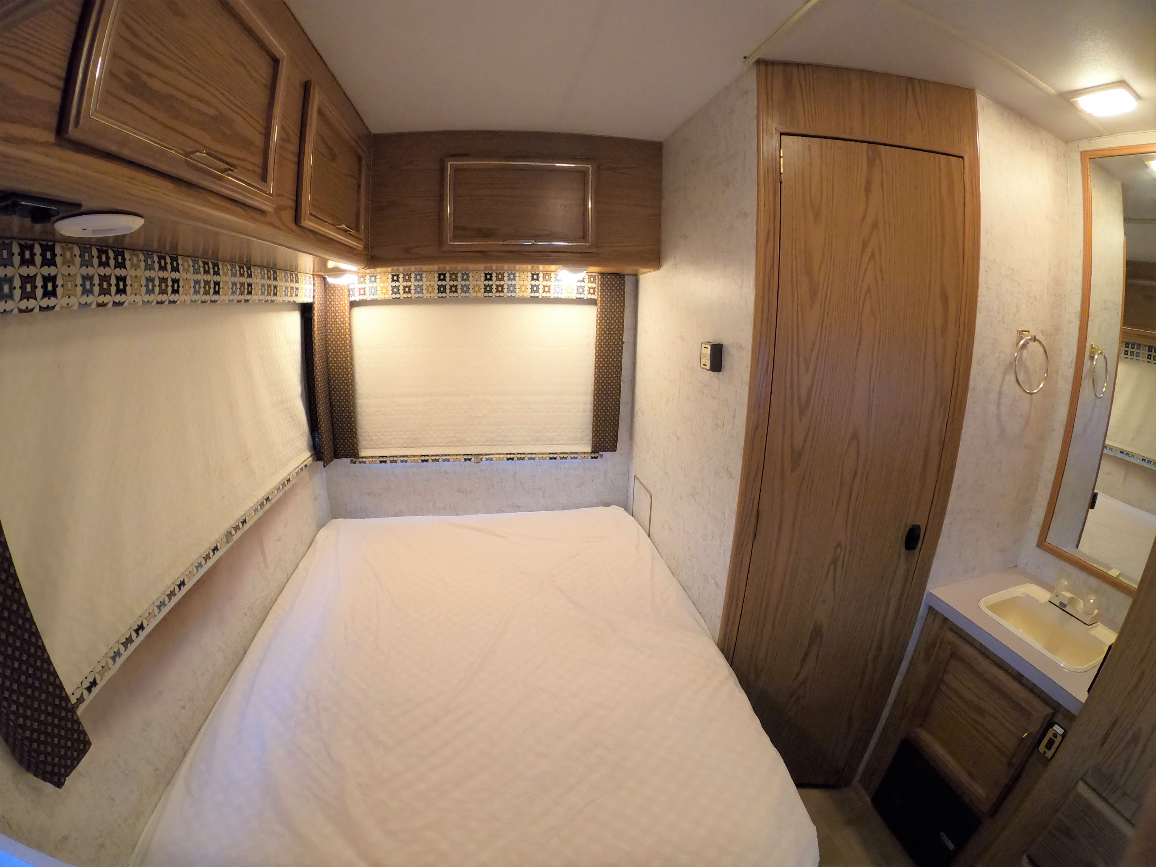 Cozy RV rental bedroom with a full bed, overhead wood cabinets, and a compact bathroom vanity.. Ford Tioga Montara 1993