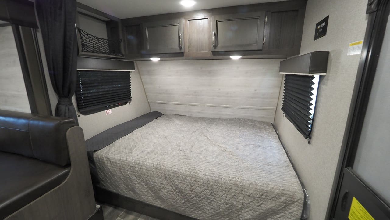 Explore this RV rental's bedroom: a comfy bed, overhead cabinets, window blinds, and a neat storage net.. Jayco 184BS 2022