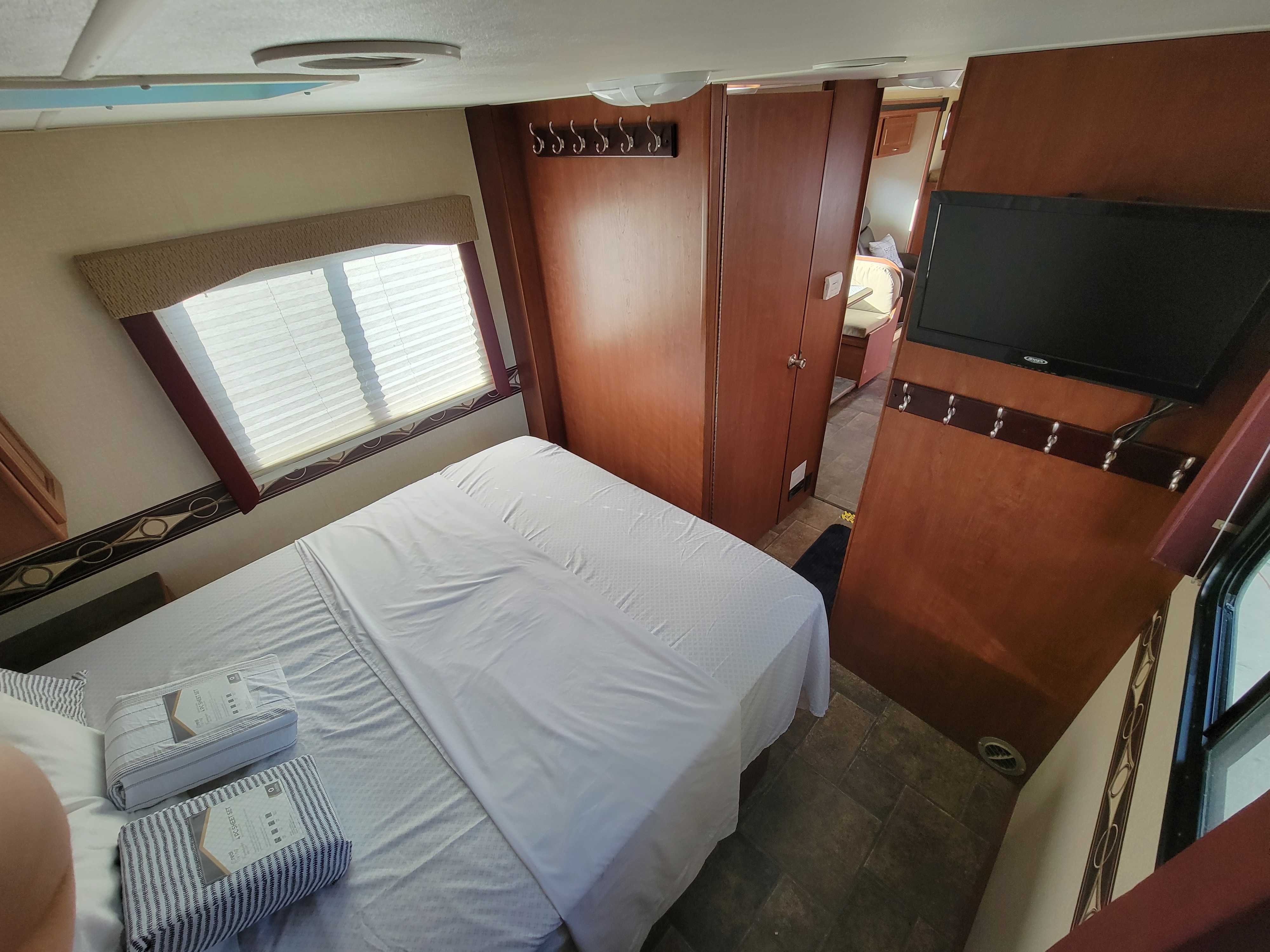 View of an RV rental bedroom with a neatly made bed, window with blinds, mounted TV, and coat hooks.. Winnebago Minnie Winnie 2014