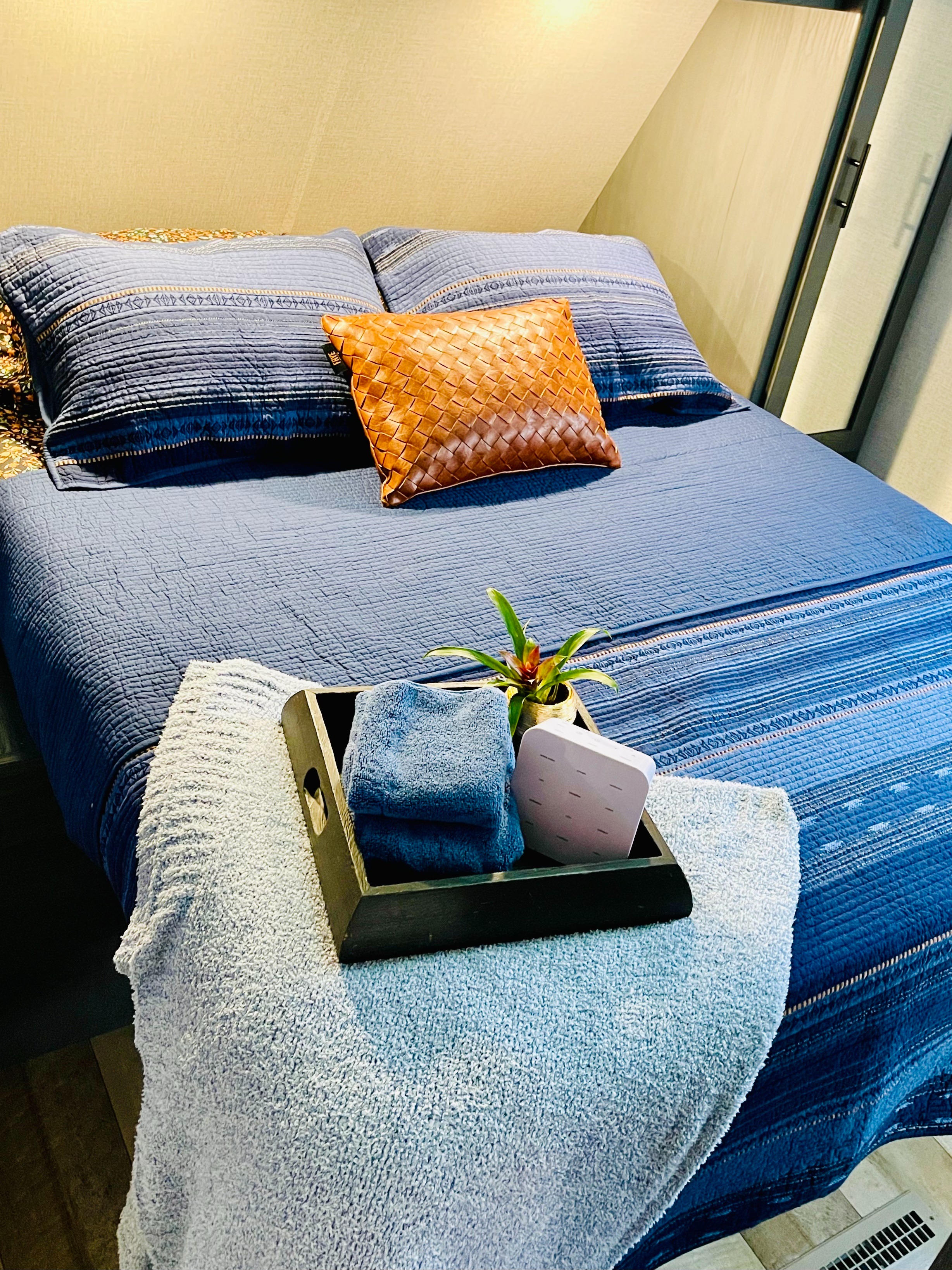 Cozy RV rental bedroom with blue bedding, accent pillows, and a welcome tray with towels and a plant.. Dutchmen Aspen Trail 2022