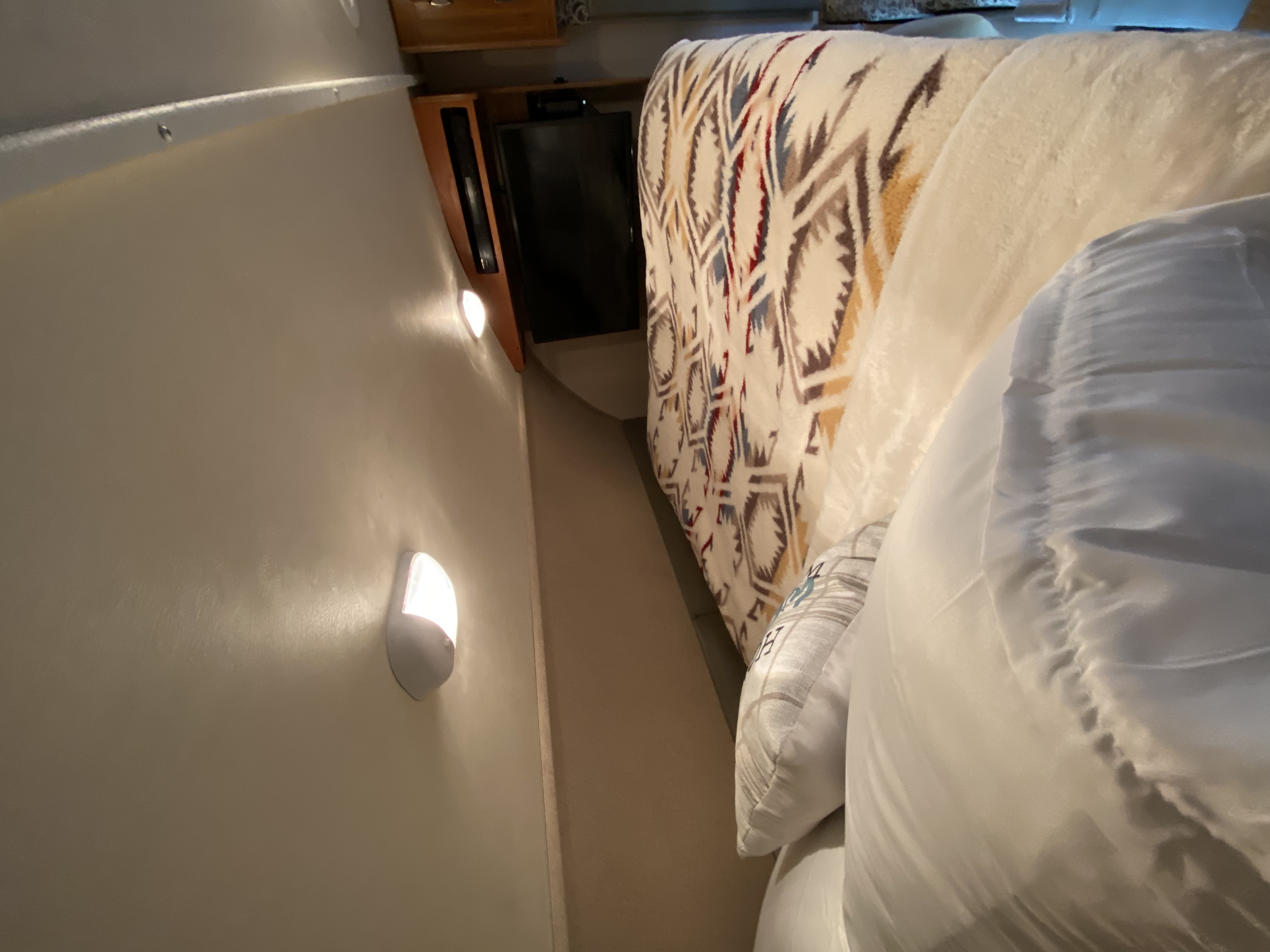 Inside an **rv rental**: a cozy bedroom with patterned blanket, white pillows, a wall-mounted TV, and bright sconce lighting.. Jayco Redhawk 2016