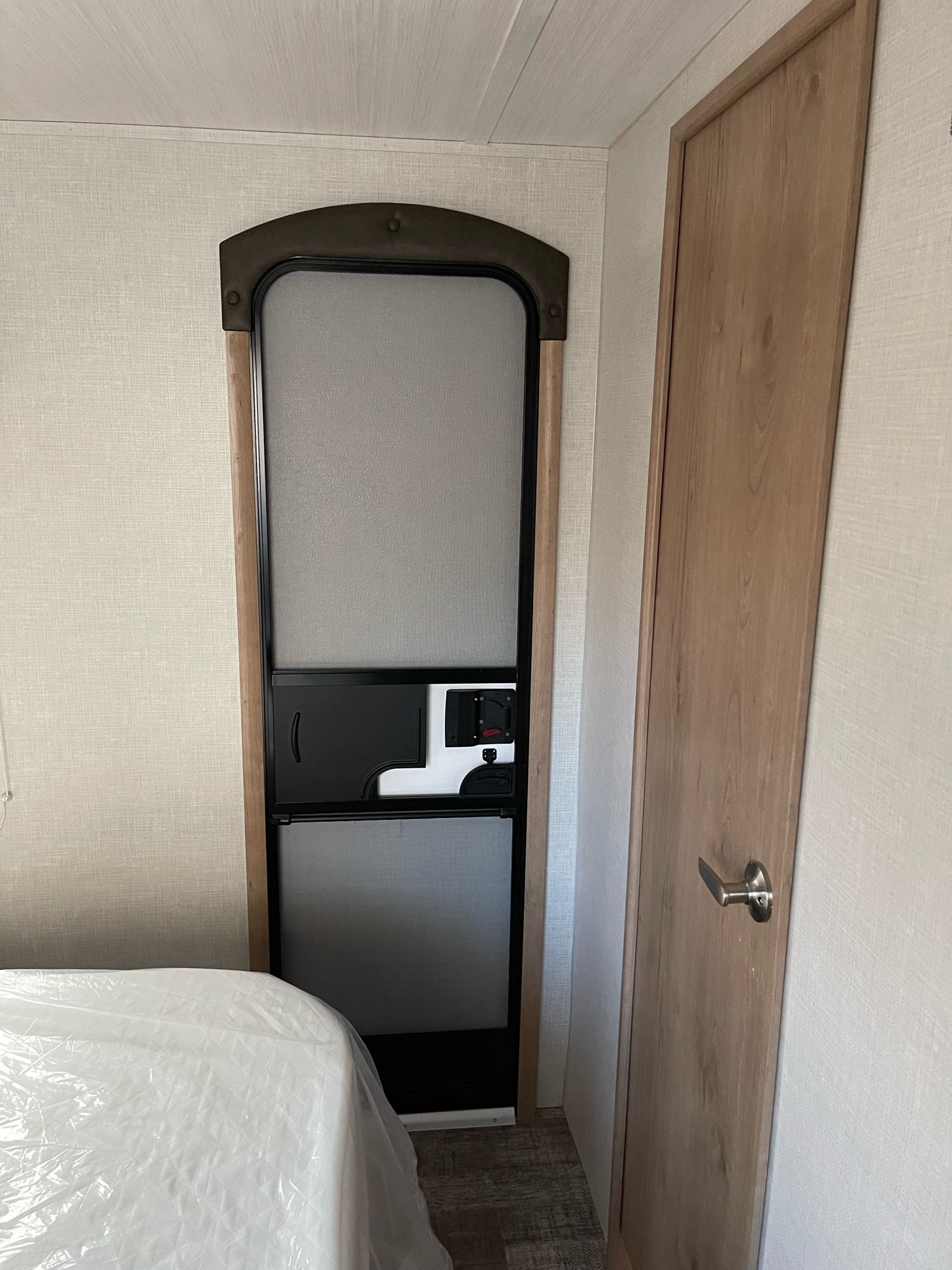 RV rental interior shows a bedroom with a bed (plastic-covered), entry door, and a light wood interior door in a travel trailer.. Winnebago Minnie 2020