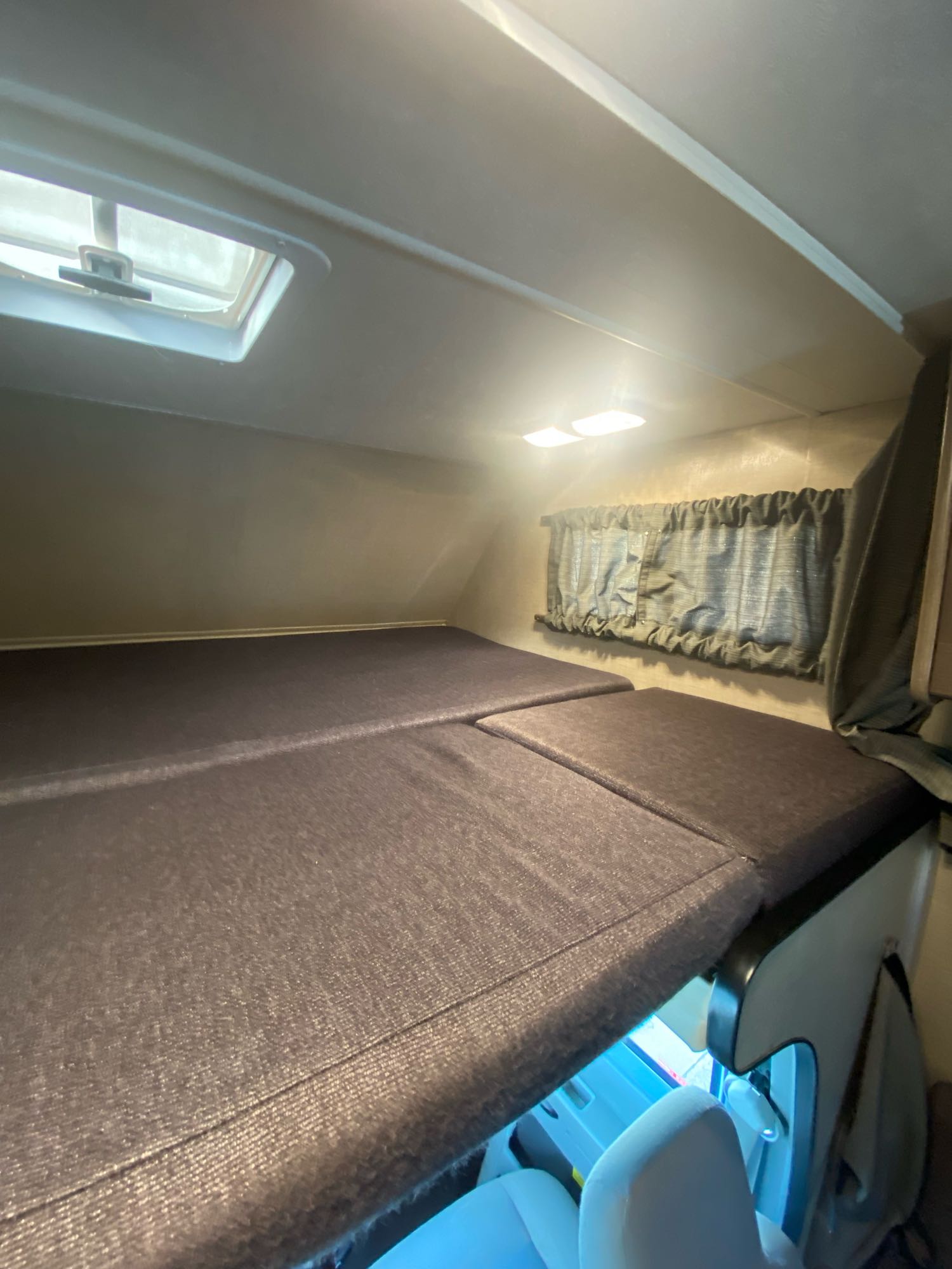 Inside an RV rental: a cozy cab-over bunk with dark mattresses, a window, overhead lights, and a roof vent.. Thor Majestic 2018