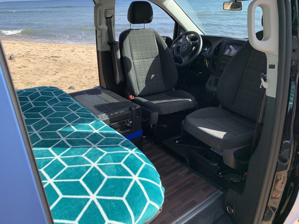 RV rental: Camper van interior featuring a comfy bed, portable fridge, and stunning beach view. Your adventure awaits!. Mercedes Getaway 2021