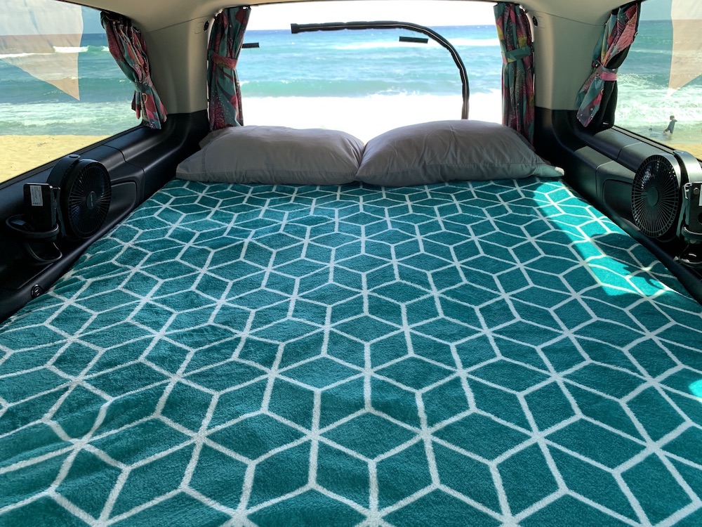 Cozy RV rental bed with pillows & teal blanket, flanked by fans, overlooks sunny beach & ocean.. Mercedes Getaway 2021