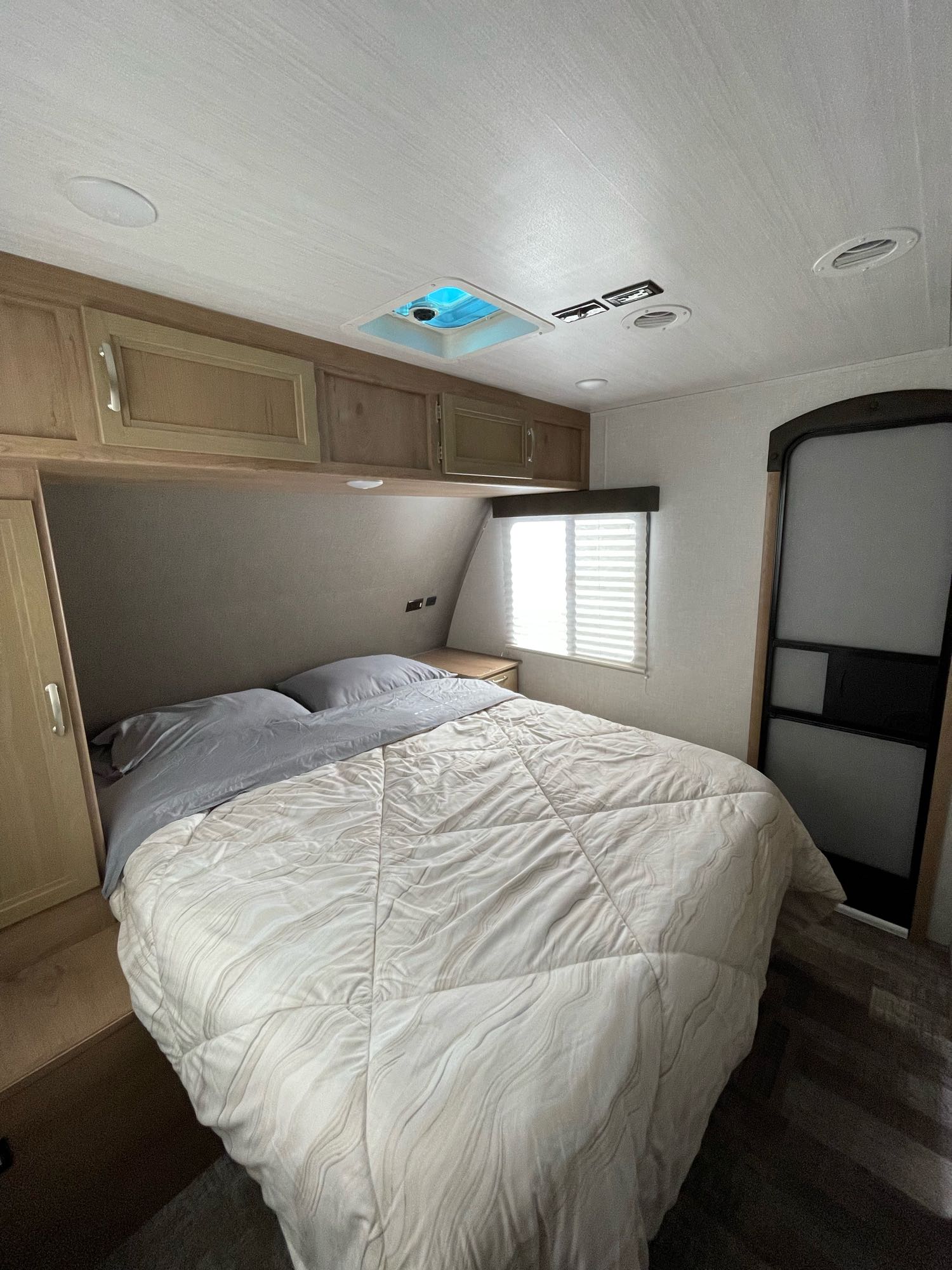 Comfortable RV rental bedroom featuring a large bed with gray pillows, overhead cabinets, and a window with blinds, ideal for travel.. Winnebago Minnie 2020