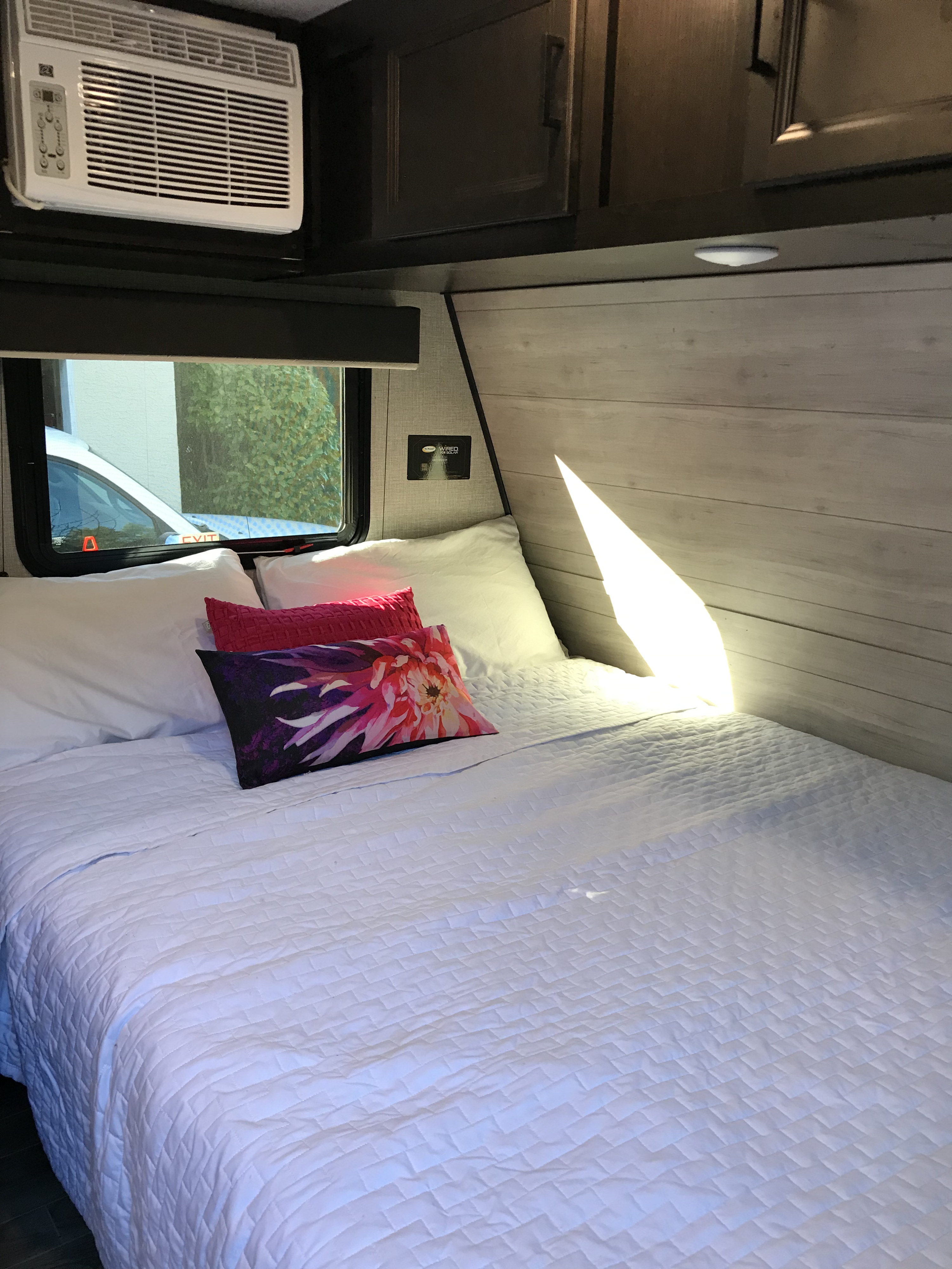 RV rental bedroom with white bed, colorful pillows, wall AC, and window. Sunbeam brightens the cozy space.. Jayco Jay Flight 2022