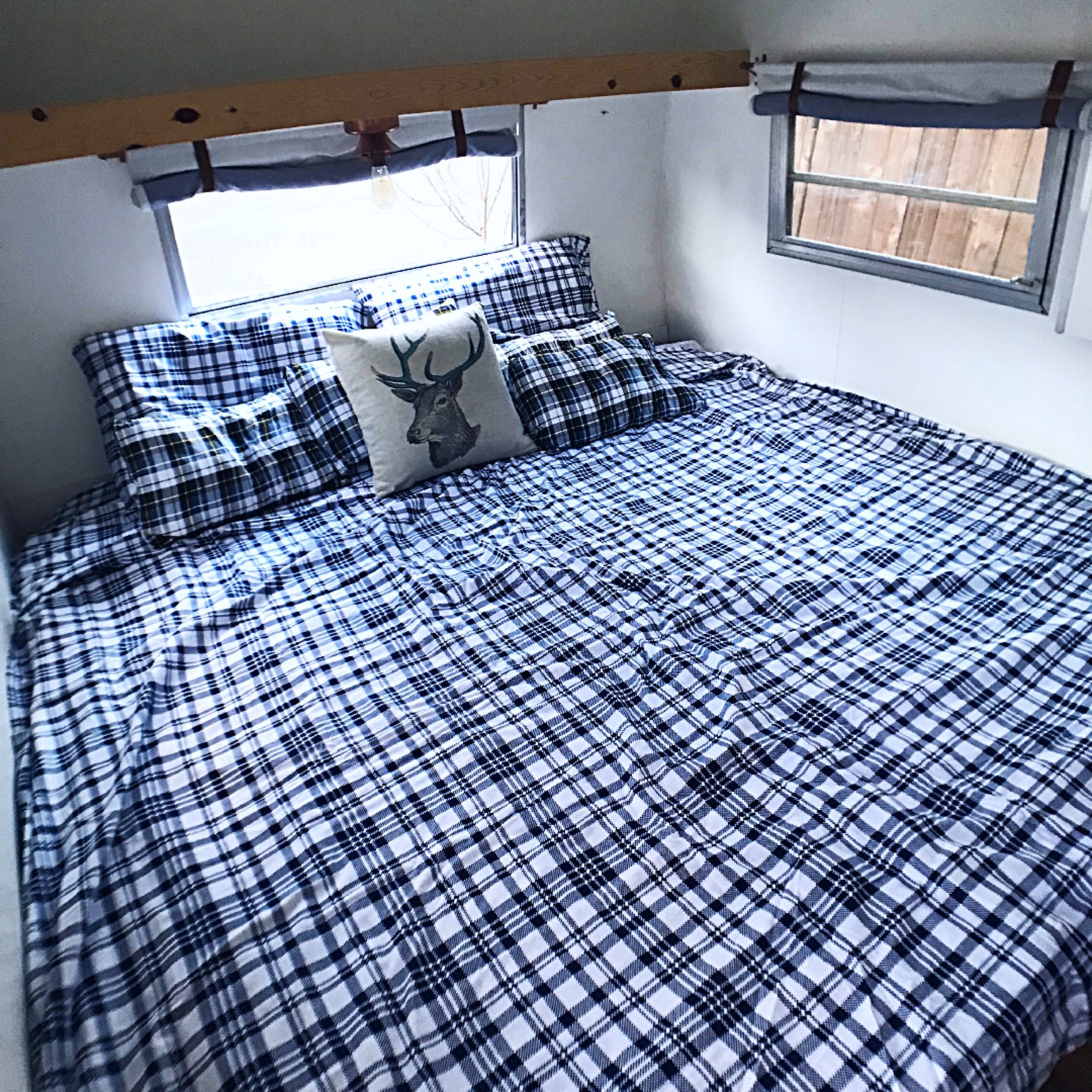 RV rental bedroom: cozy plaid bed, deer pillow, and bright windows inside a camper.. Shasta Compact 1971