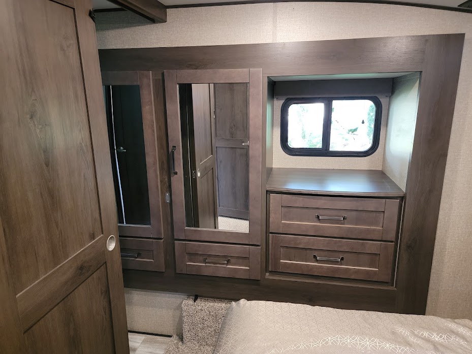 Inside an RV rental bedroom featuring custom wood cabinetry, built-in dresser, mirrored wardrobes, and a small window.. Grand Design Reflection 2020