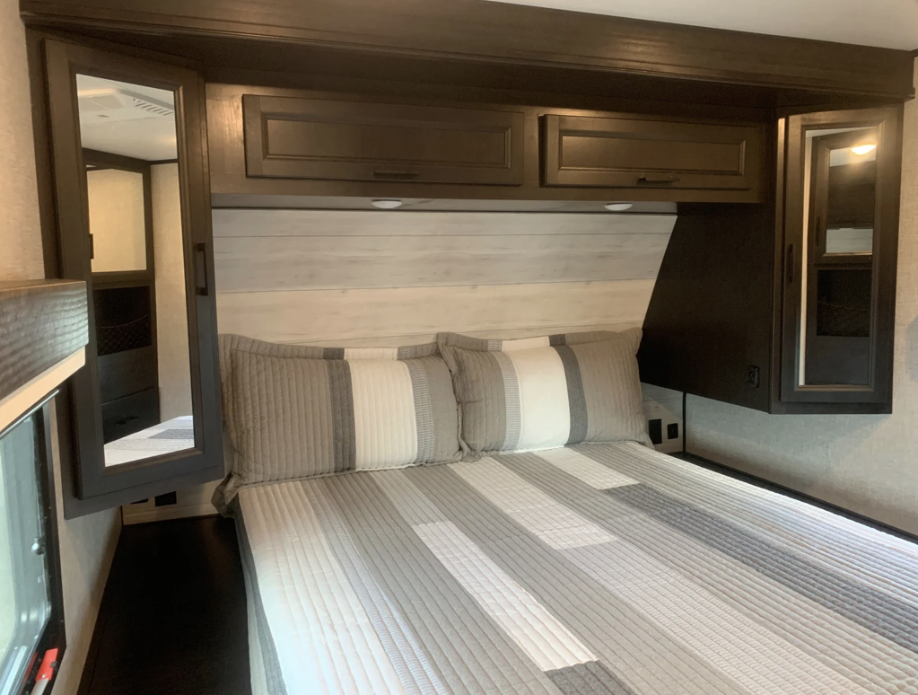 View of an RV rental bedroom with a comfy bed, striped bedding, dark wood cabinets, and mirrored storage on each side.. Jayco EAGLE 2022